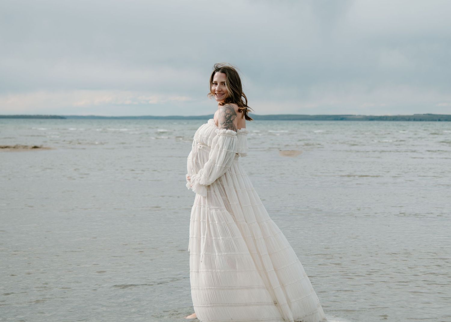Elegant maternity beach photo with flowing white dress against misty coastal backdrop. Calgary Alberta Maternity Photographer.