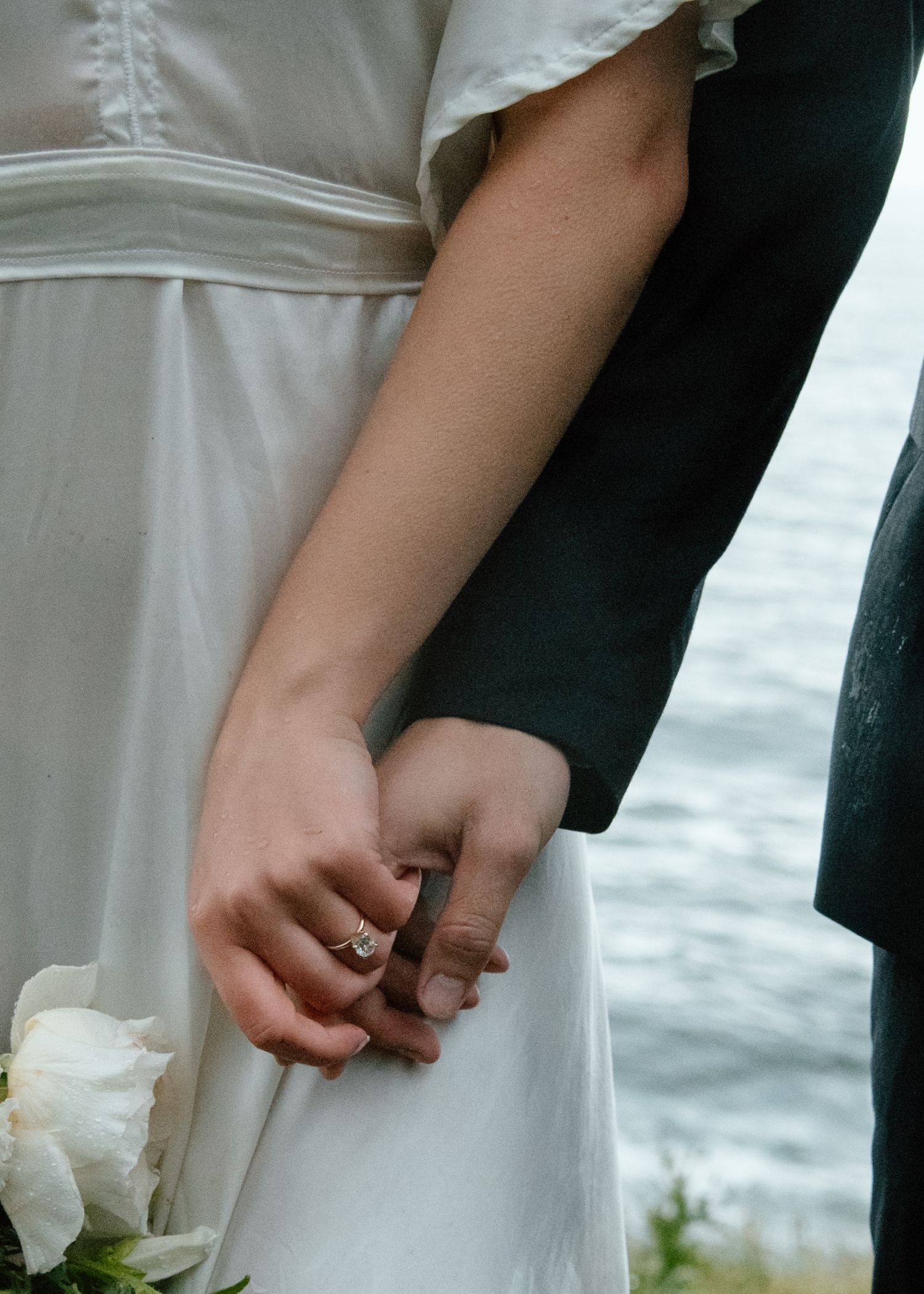 Close-up of joined hands showing wedding rings against a white dress and ocean backdrop. Calgary Wedding Photographer