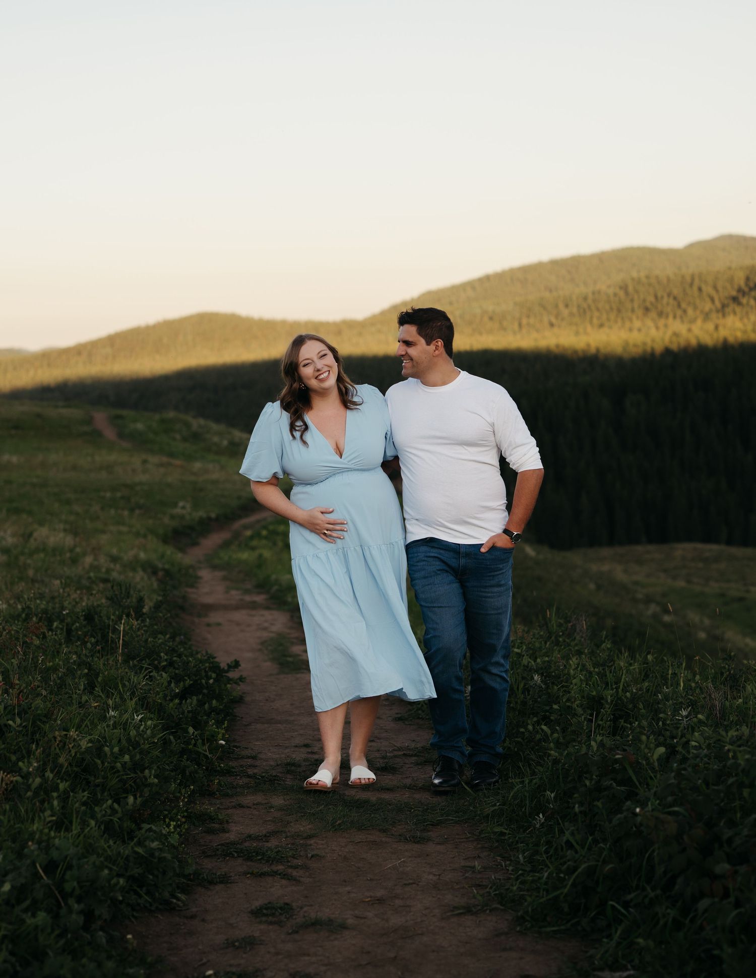 A couple stands on a scenic dirt path at sunset with rolling hills in the distance creating a romantic silhouette.