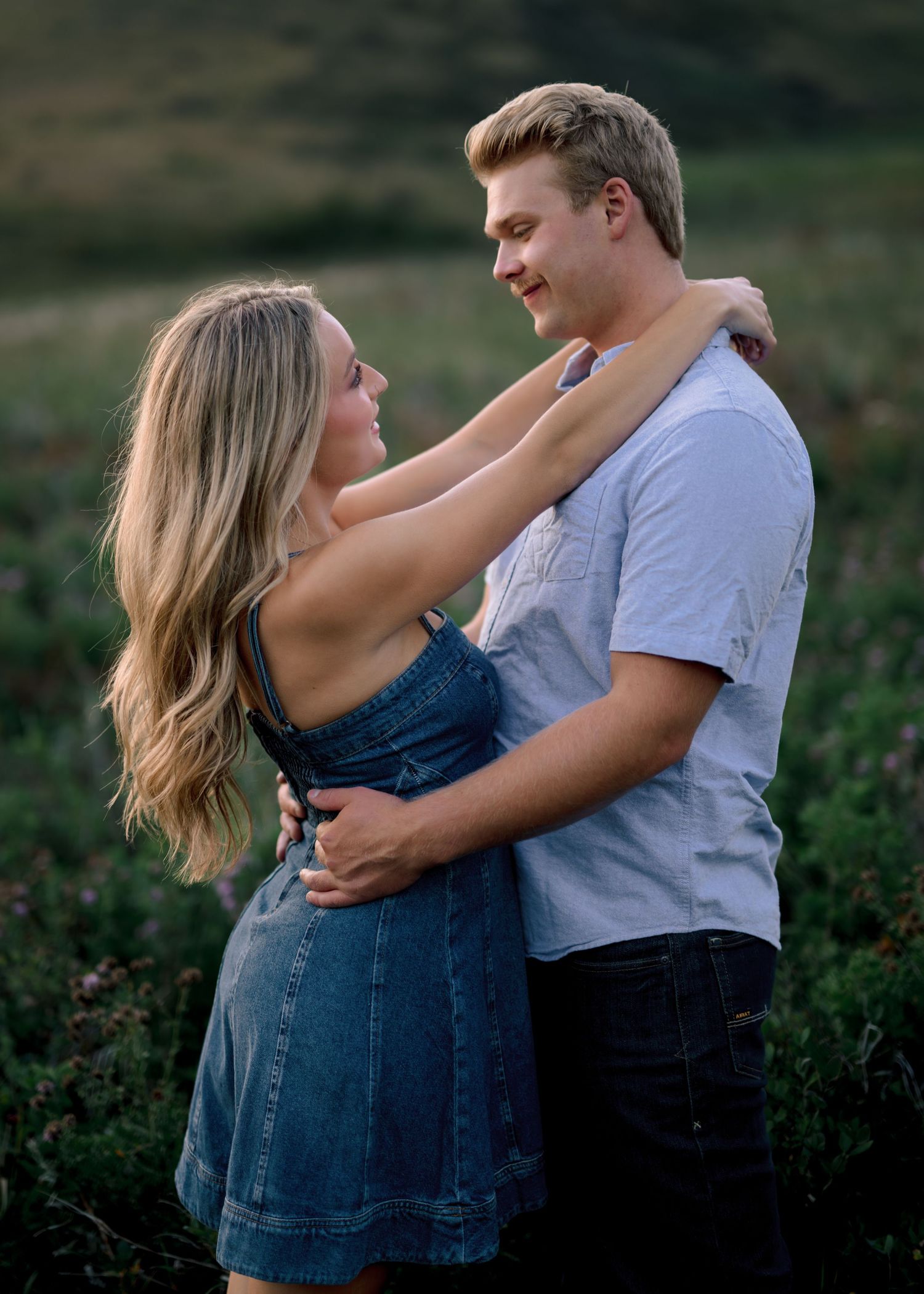 A couple embraces outdoors in casual clothing during sunset with blurred greenery in the background.