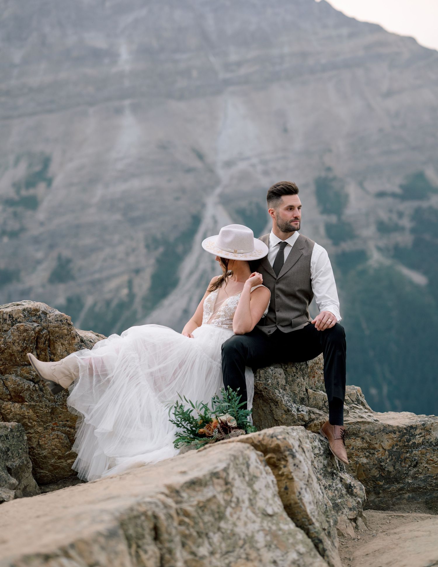 A couple in wedding attire sits intimately on rocky mountain ledge with dramatic peaks in background.