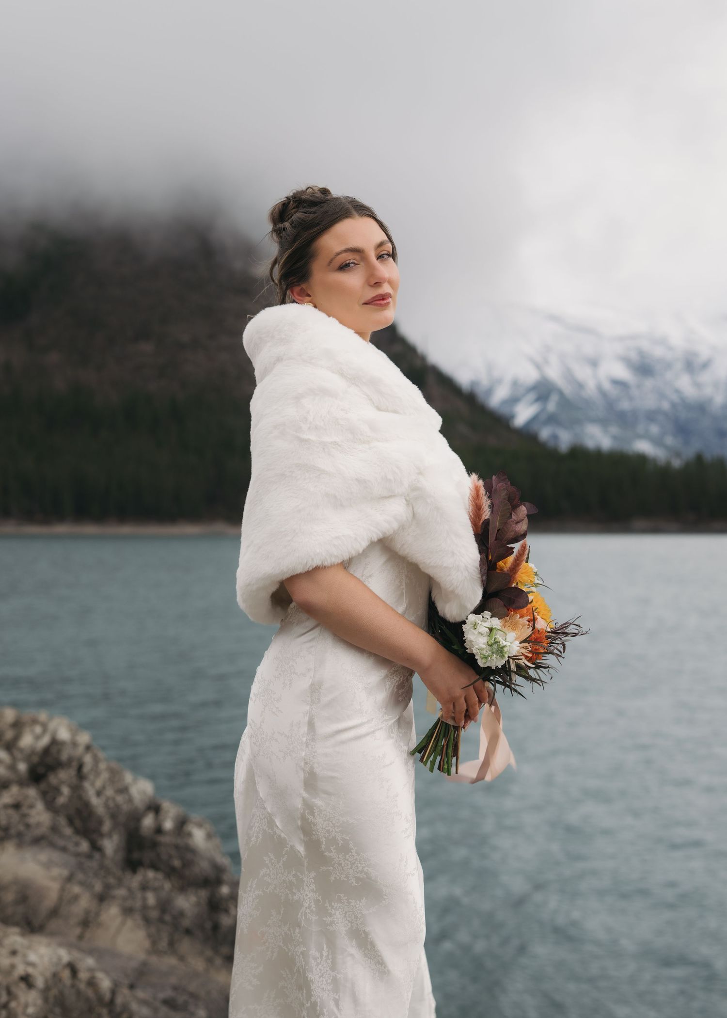 A person in a white fur wrap holds a colorful bouquet against a snowy mountain and lake backdrop.