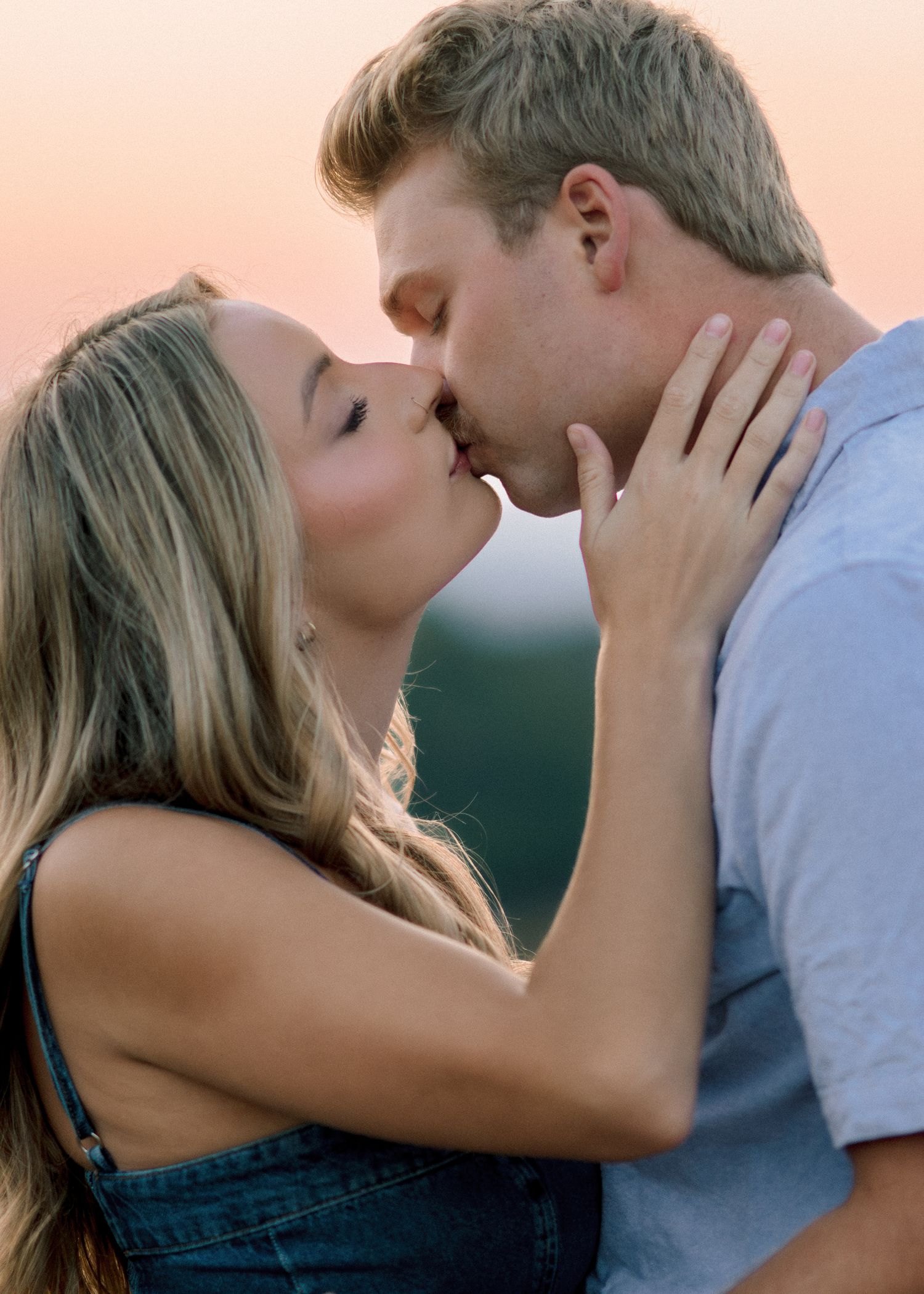 A romantic couple sharing a tender kiss at sunset against a soft rosy backdrop.