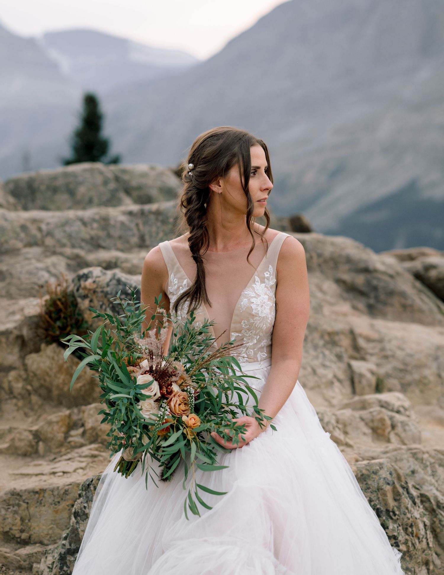 A delicate white wedding dress and rustic bouquet set against a scenic mountain landscape and rocky terrain.