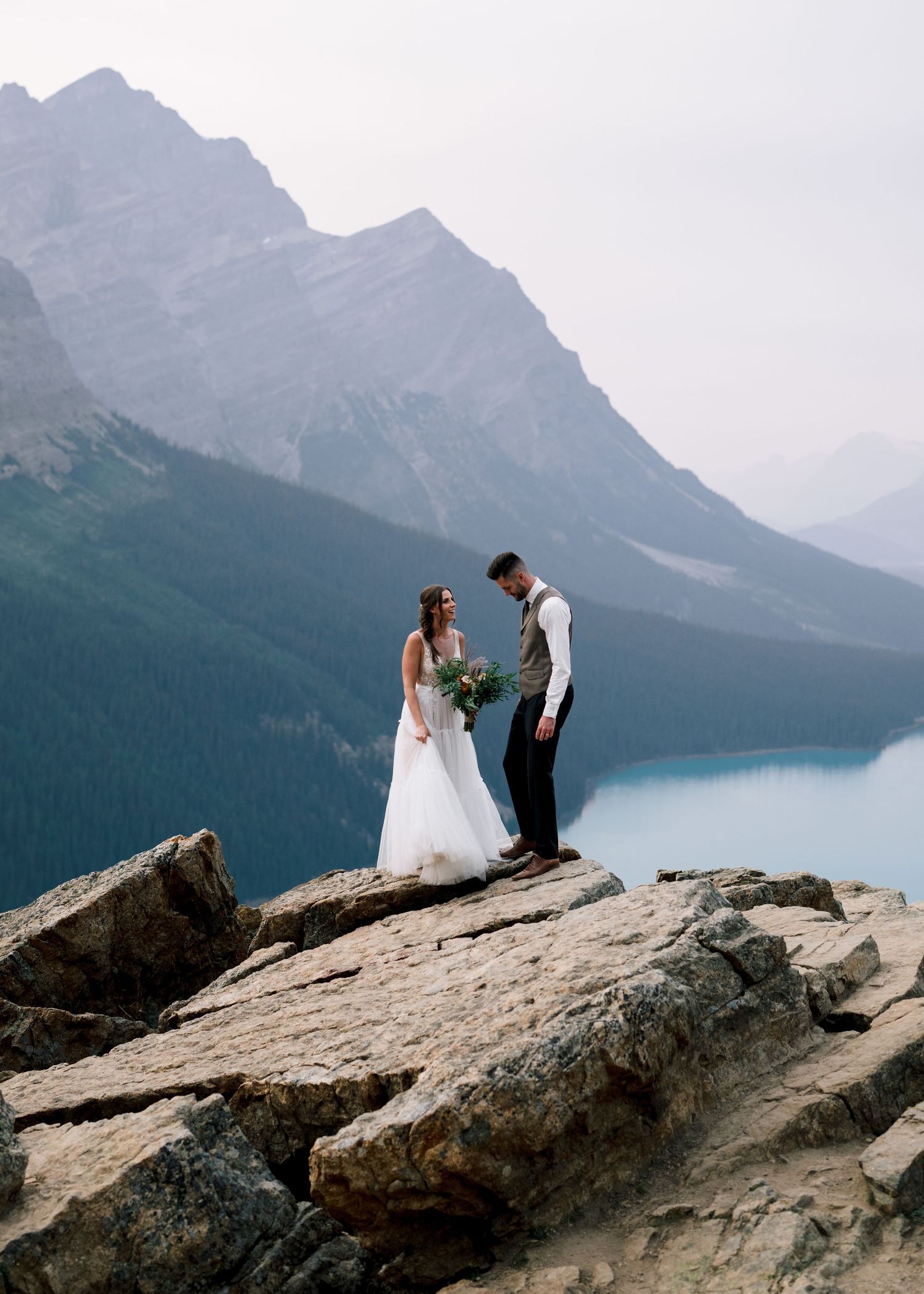 A couple in wedding attire stands on rocky cliffs overlooking a misty mountain lake with dramatic peaks in the background.