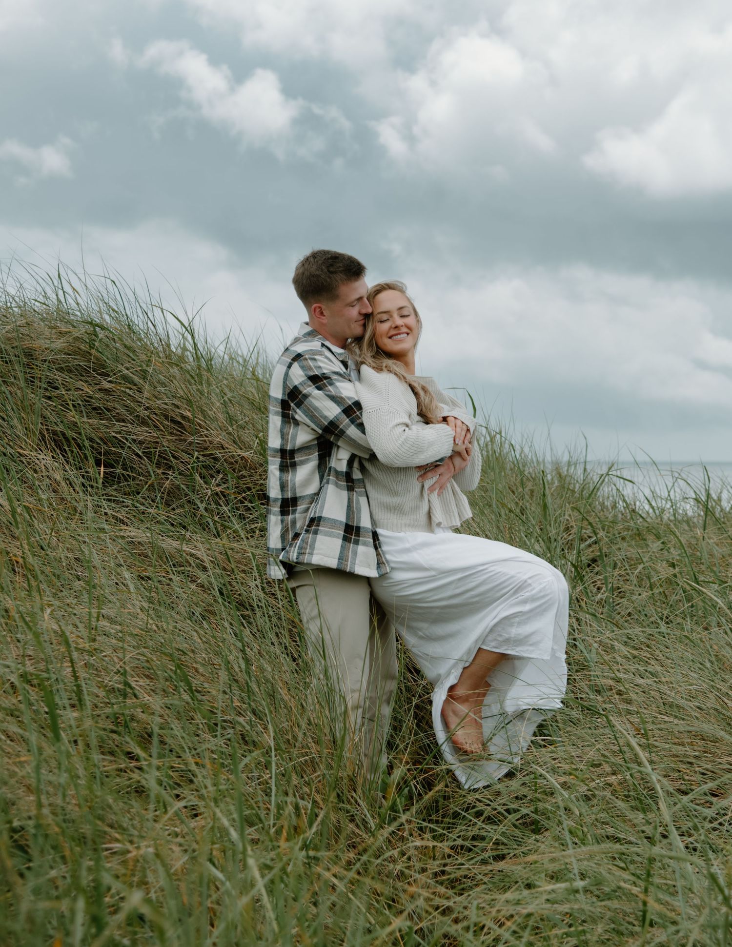 Romantic couple embraces on grassy hillside with moody cloudy sky backdrop on Oregon Coast. Calgary Lifestyle and Engagement Photographer.