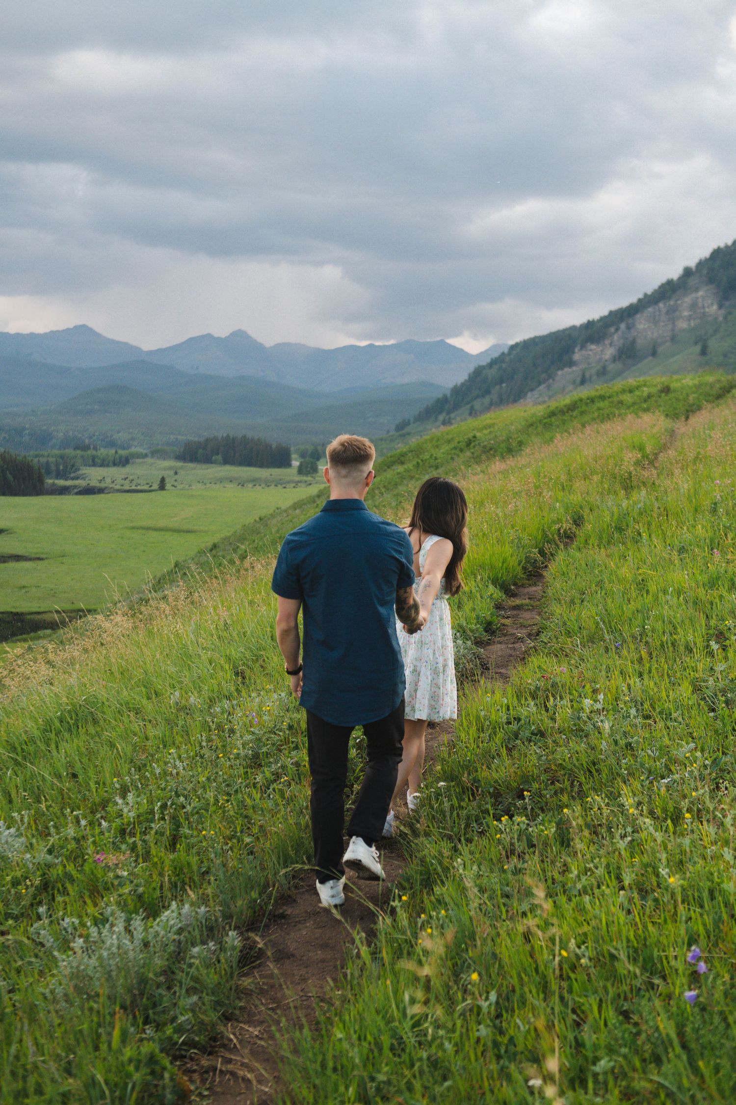Two people hike along a mountain trail through wildflower meadows with scenic mountain views in Calgary. Calgary Lifestyle and Engagement.