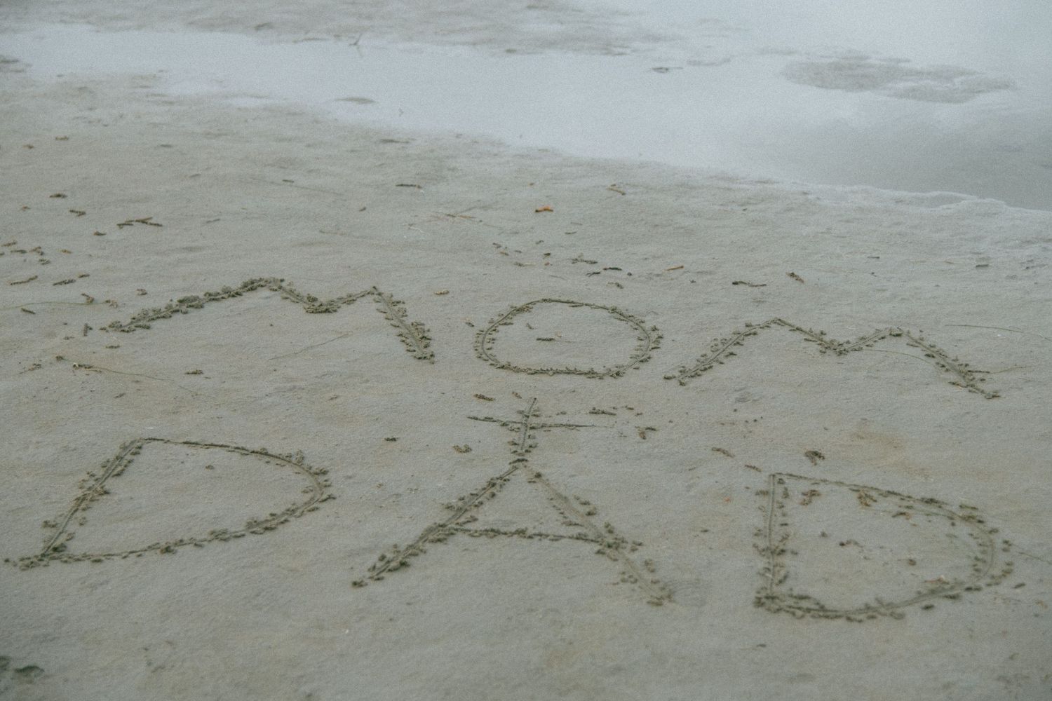 Text written in sandy beach reads 'MOM DAD' with ocean waves in the background. Calgary Alberta Maternity Photographer.