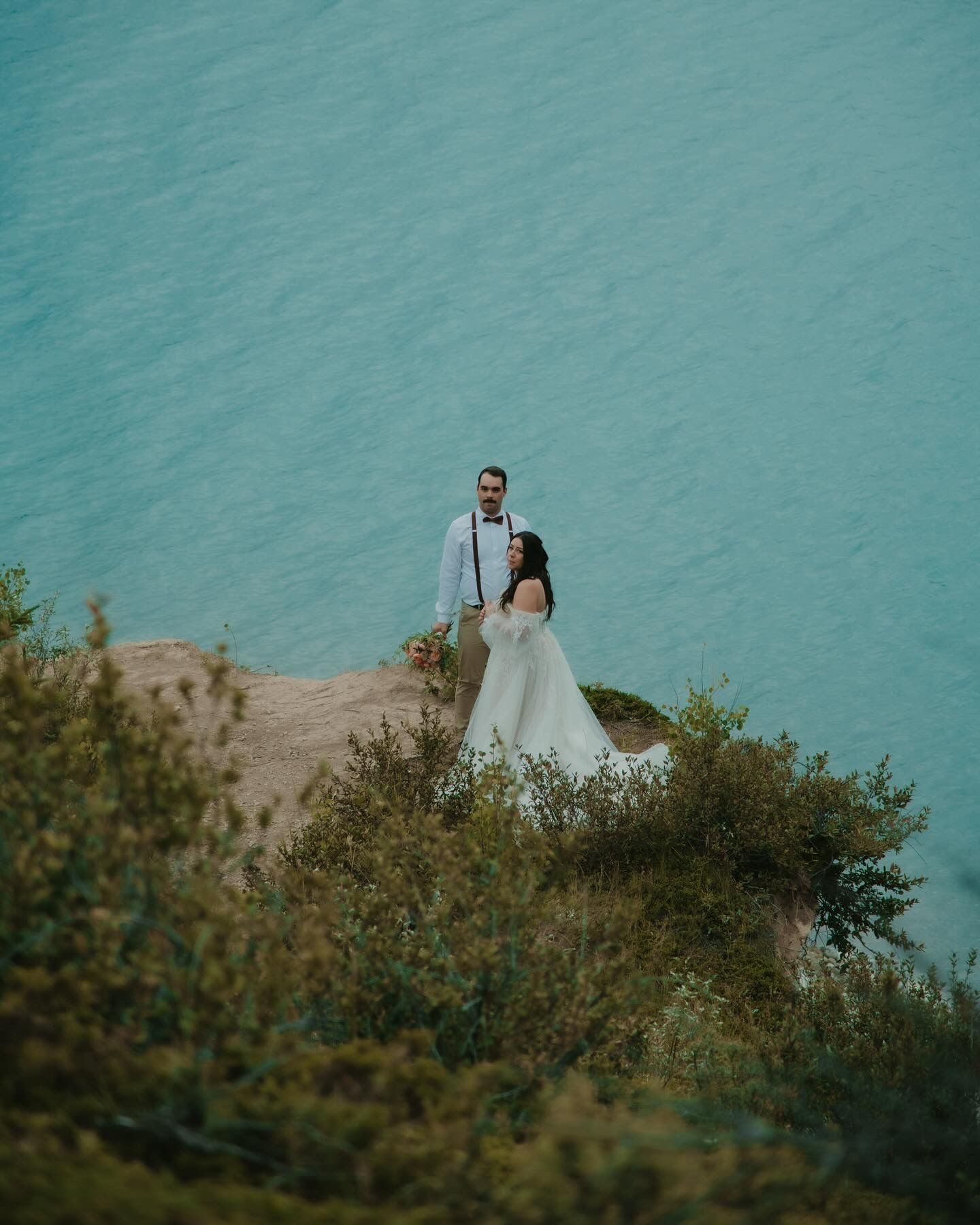 A wedding couple stands on a cliff overlooking turquoise waters. Calgary Wedding Photographer