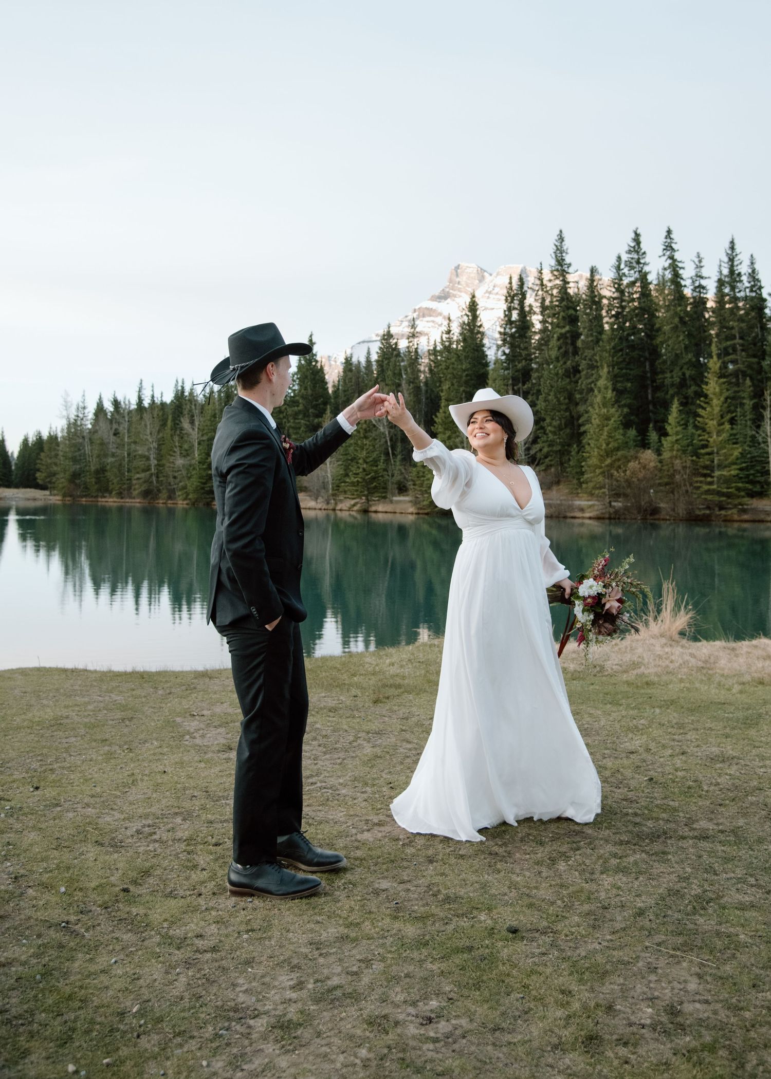 A couple in western wedding attire dances by a mountain lake surrounded by pine trees. Banff and Calgary Wedding Photographer