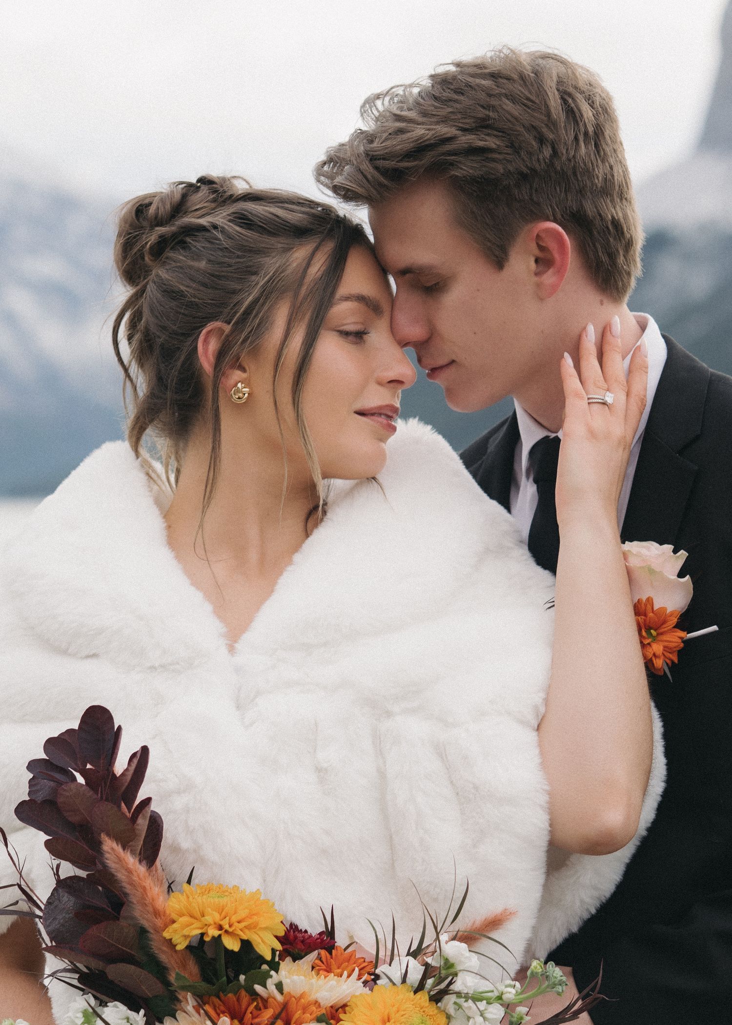 Romantic winter wedding couple embraces outdoors with burgundy and orange fall bouquet against snowy mountain backdrop.