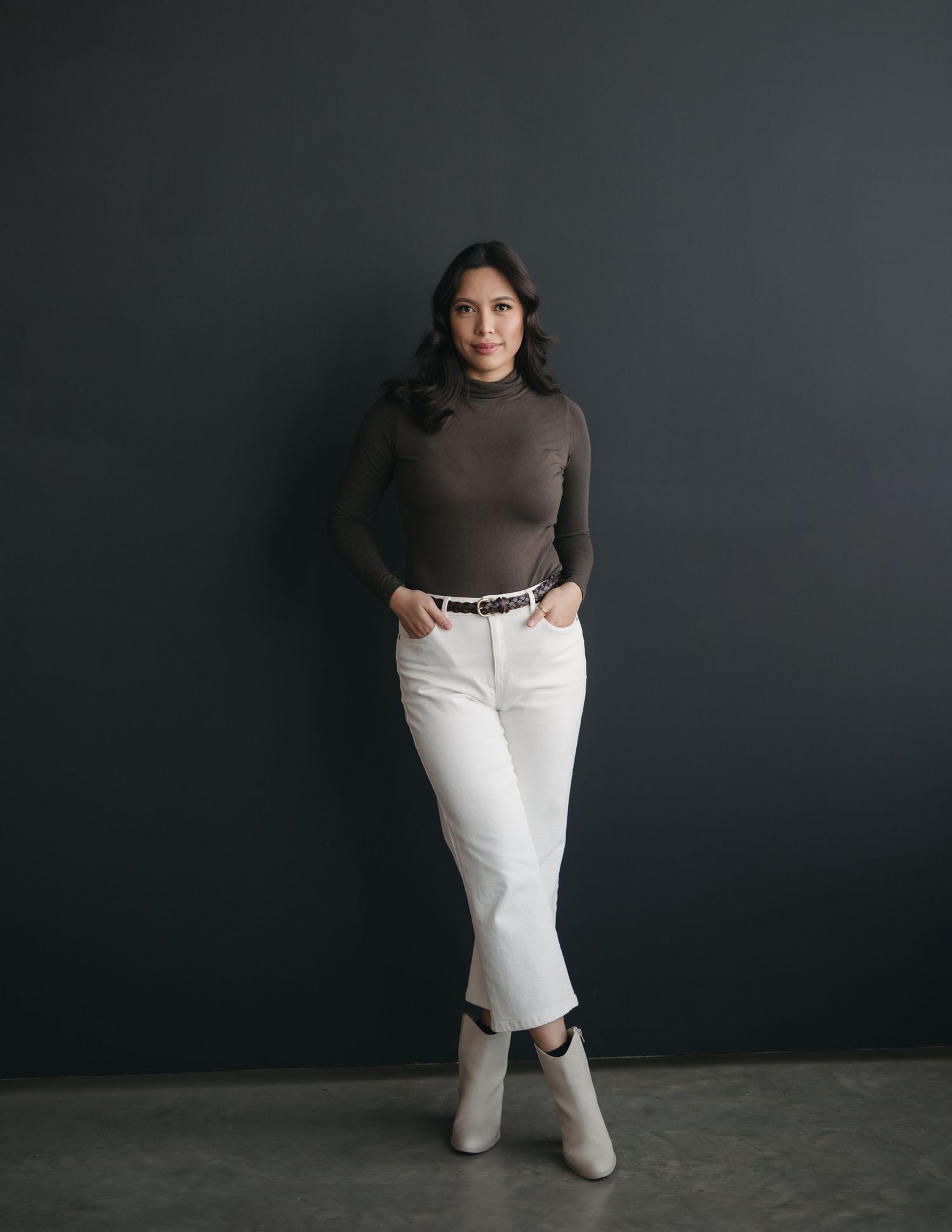 Model in dark turtleneck and white pants poses against gray backdrop in fashion studio photoshoot series.