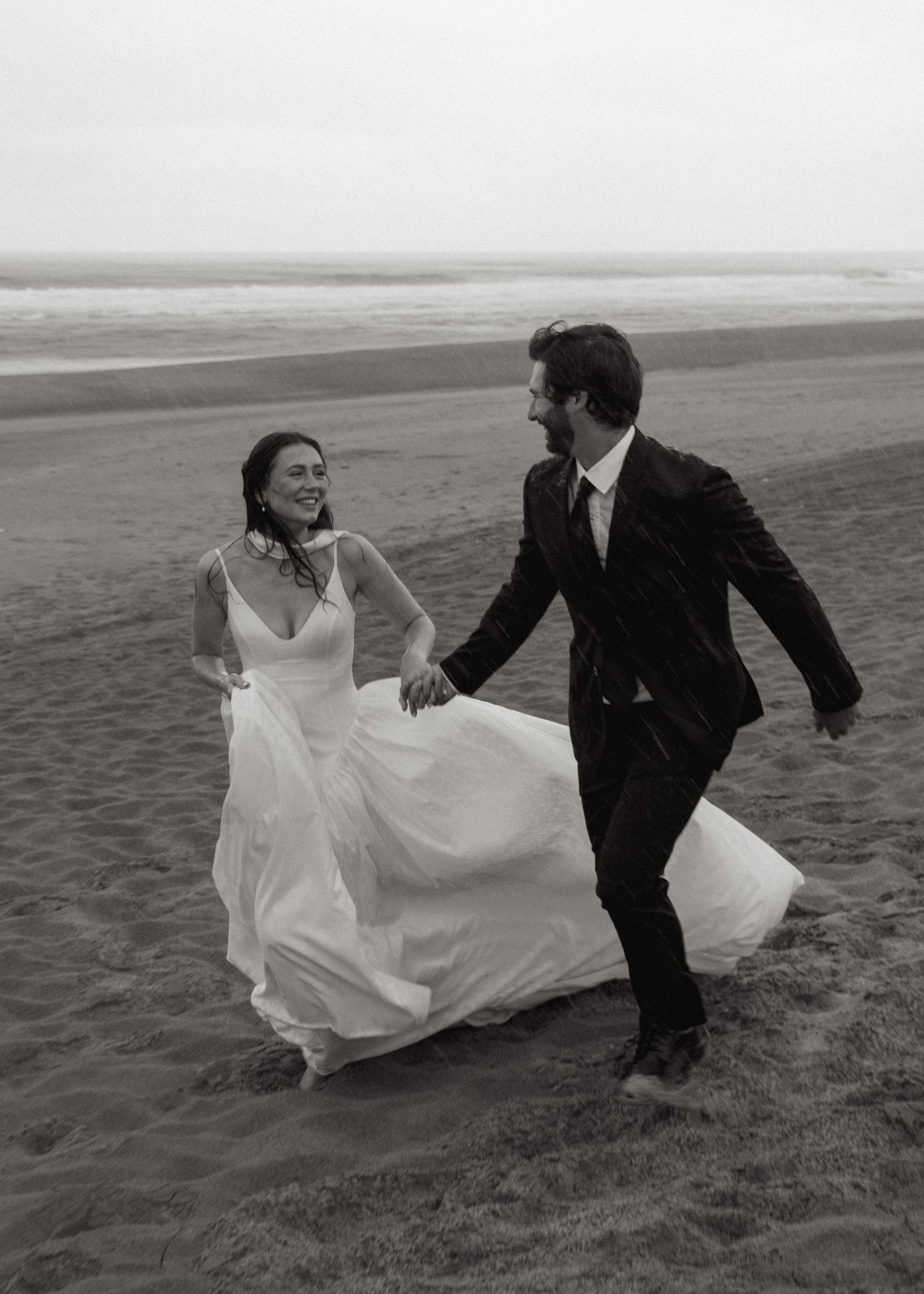 A couple runs across a sandy beach together in black and white wedding attire on their special day.