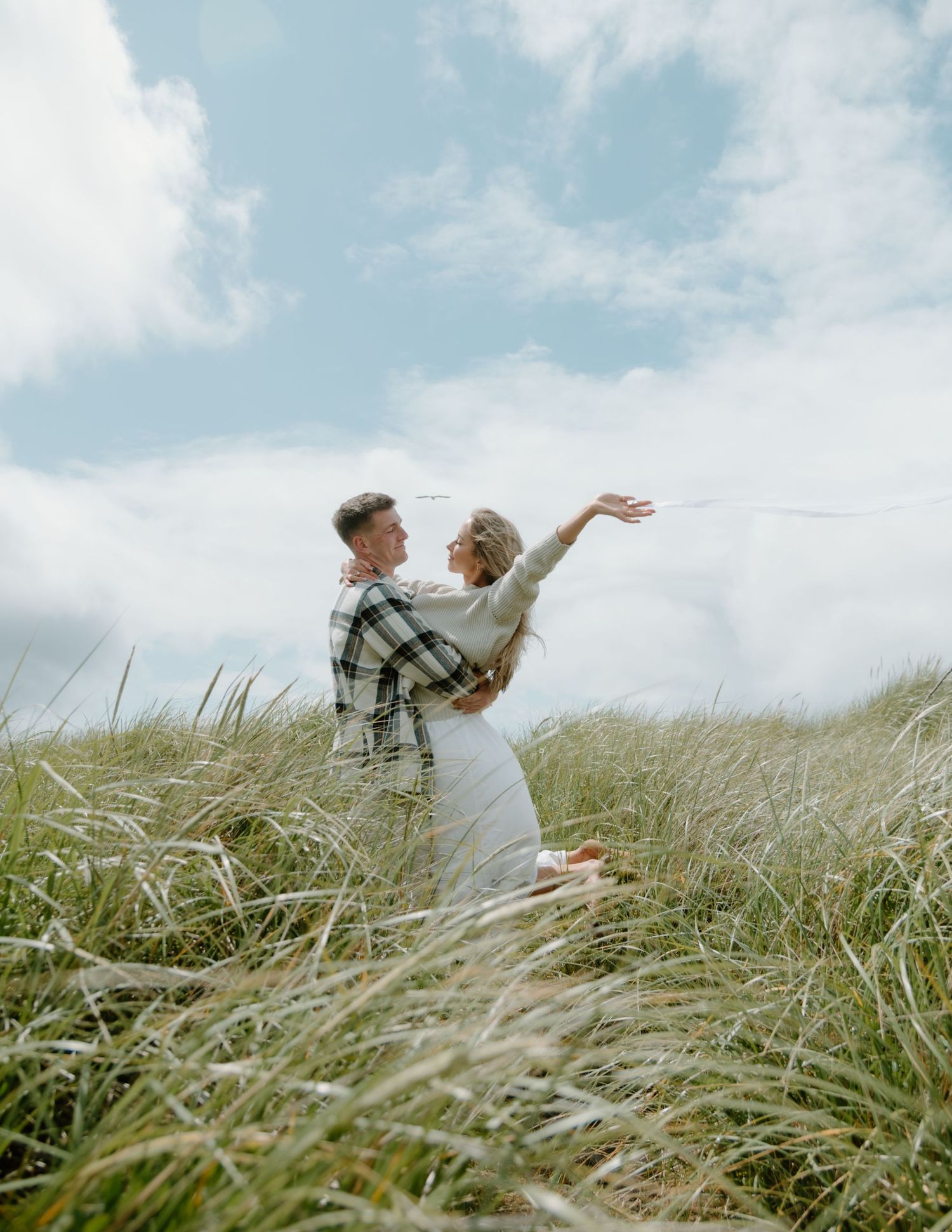 Dancing couple in white clothes twirl among windswept coastal grass on Oregon Coast. Calgary Lifestyle and Engagement Photographer.