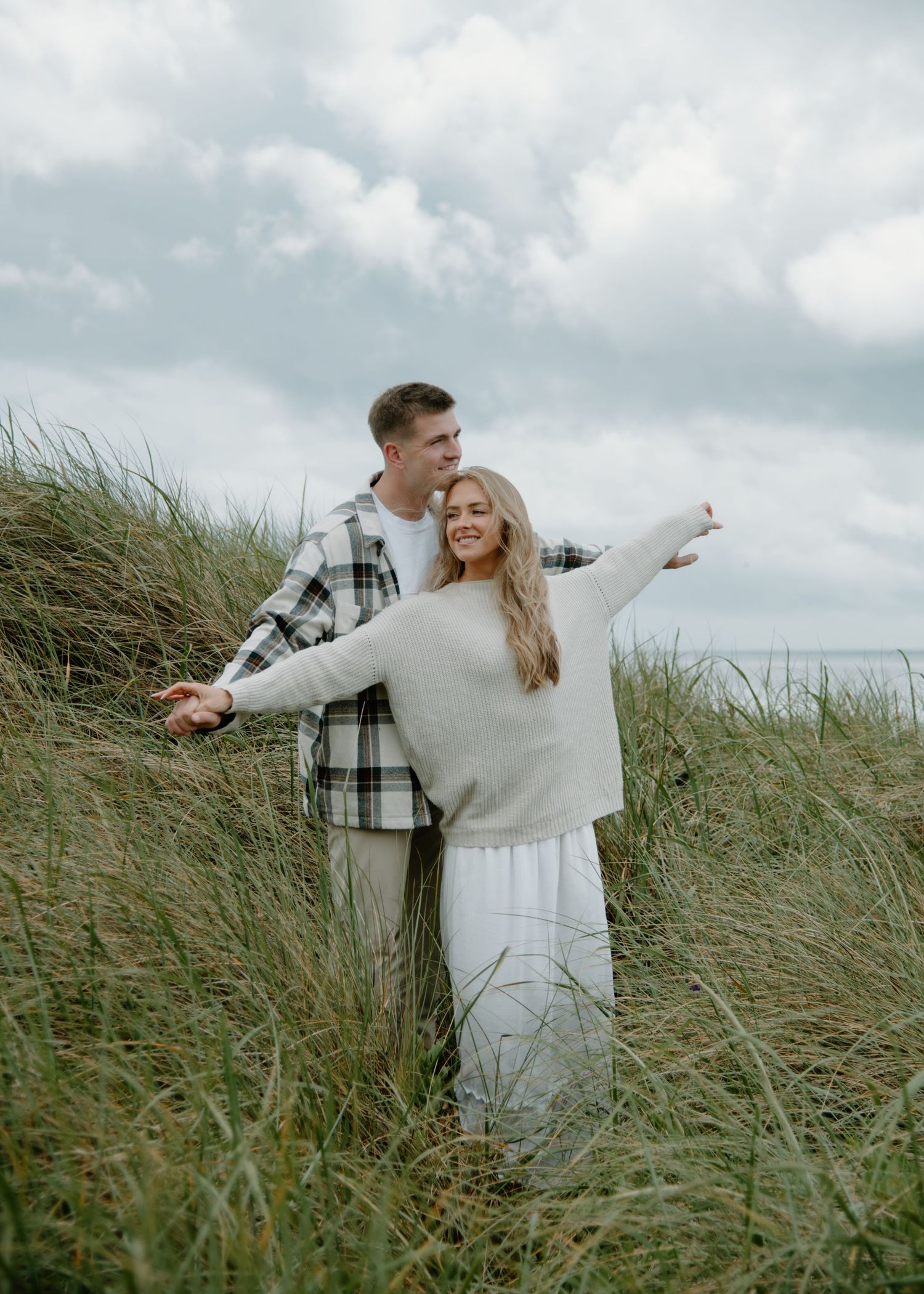 Couple stands with outstretched arms among tall grass on coastal dunes. Calgary Alberta Engagement Photographer.