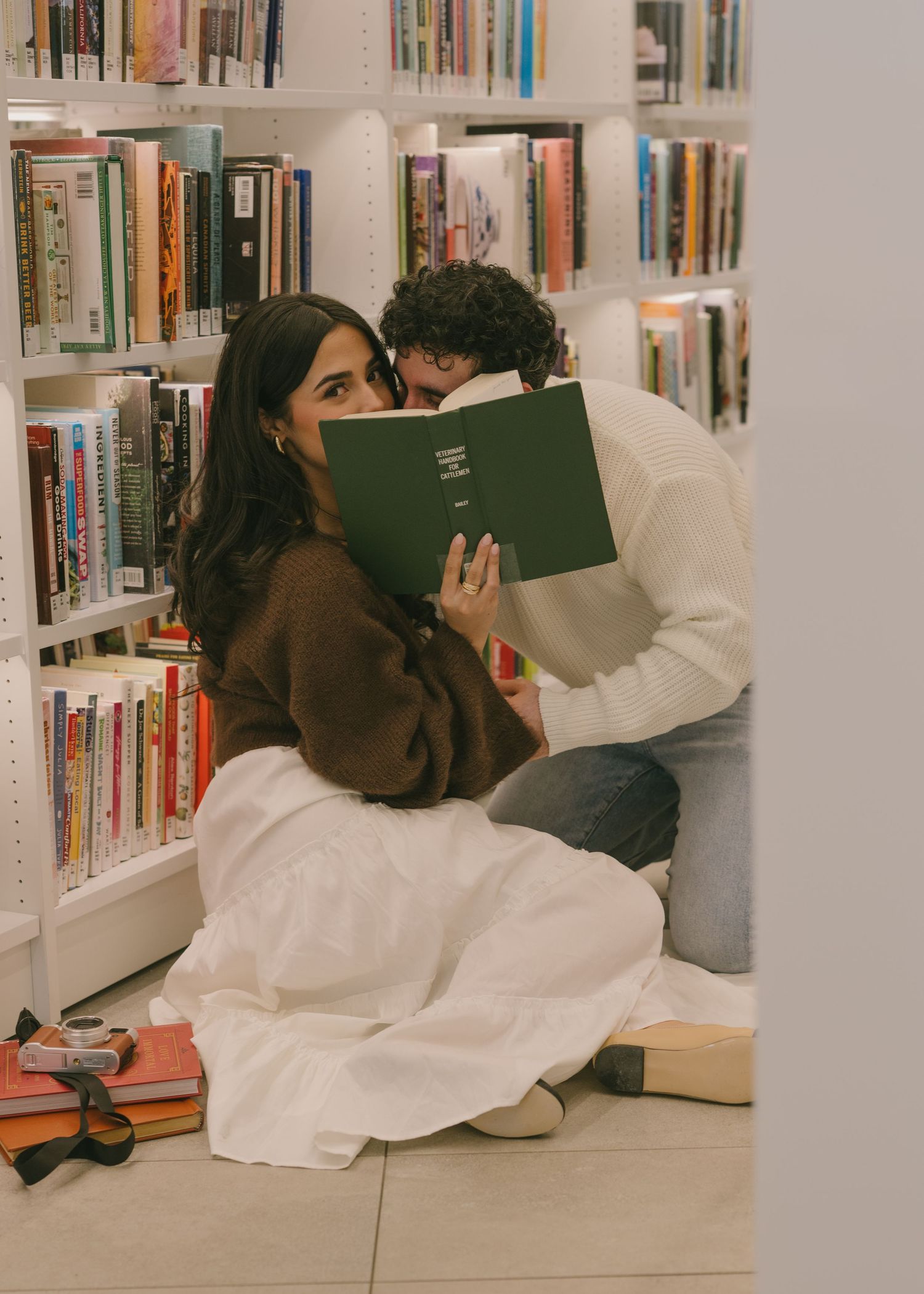 A person in a white dress lies on the floor reading a book in a library between bookshelves.