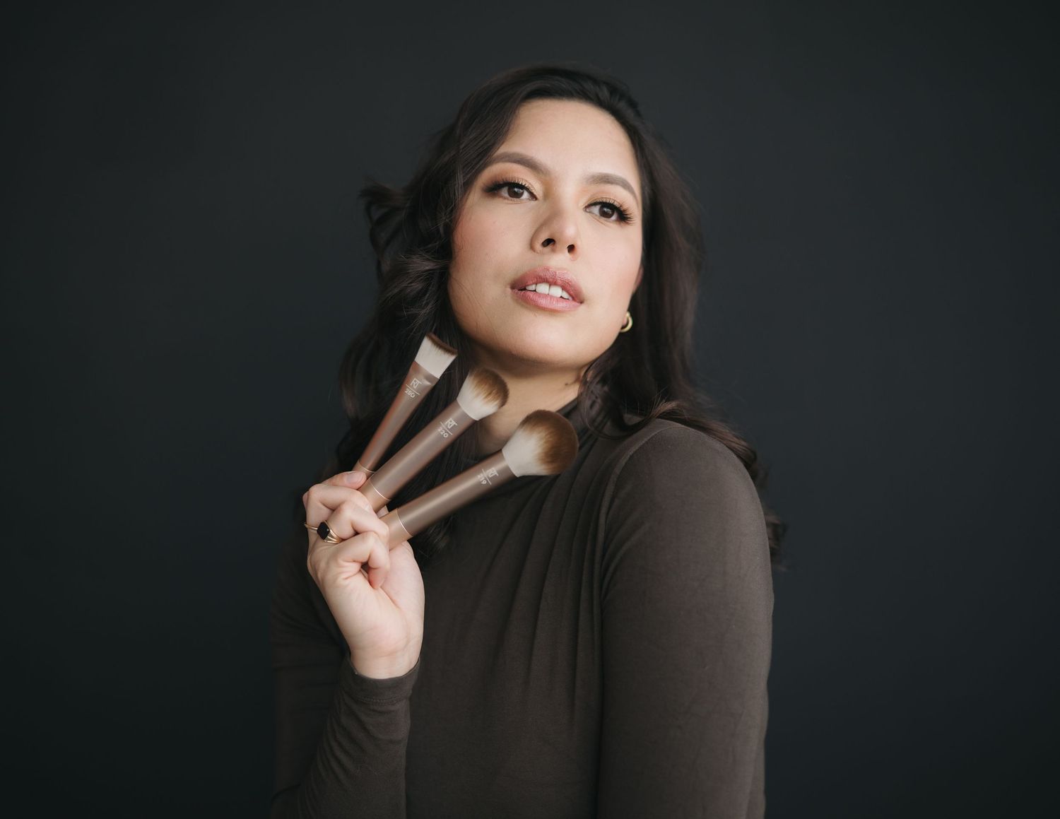 Professional makeup artist holds three cosmetic brushes against dark background in elegant portrait pose.