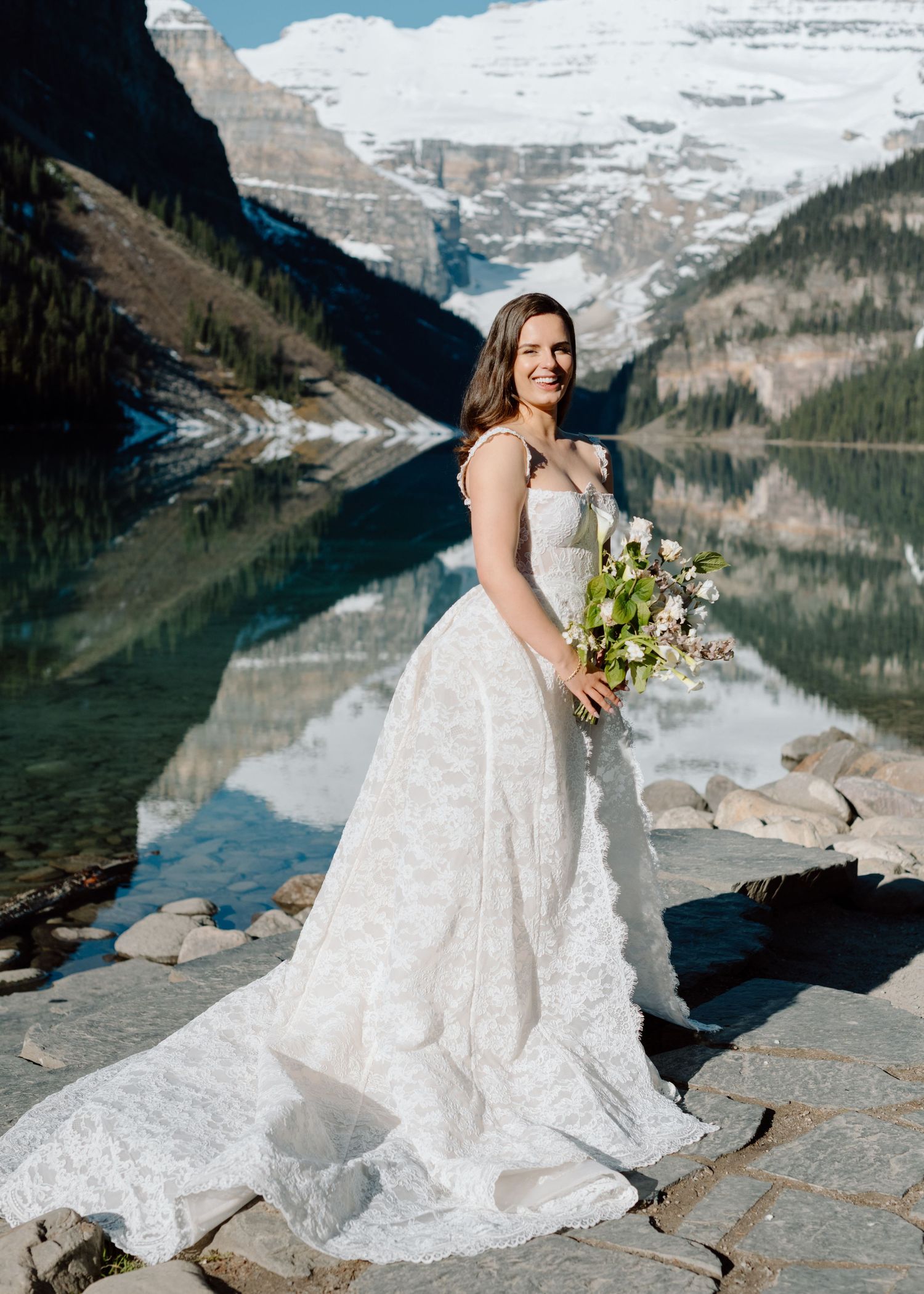 Bride in lace gown poses with bouquet at Lake Louise with snow-capped mountains in background. Banff and Calgary Wedding Photographer