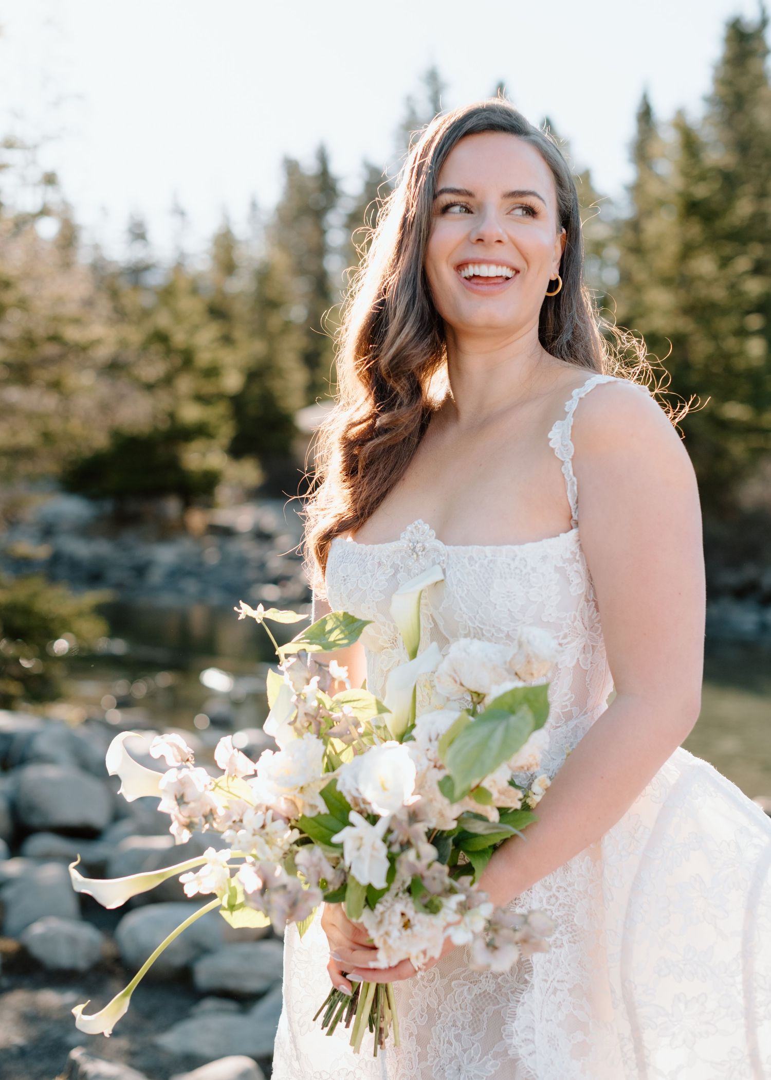 A bride in a lace dress holds a bouquet of white flowers against a rocky outdoor setting at Lake Louise. Calgary Wedding Photographer