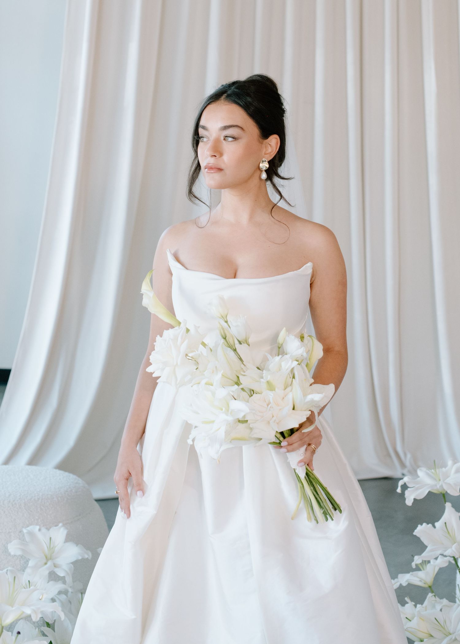 Elegant bride in white gown poses with orchid bouquet against white draped backdrop at the Brownstone YYC. Calgary Wedding Photographer
