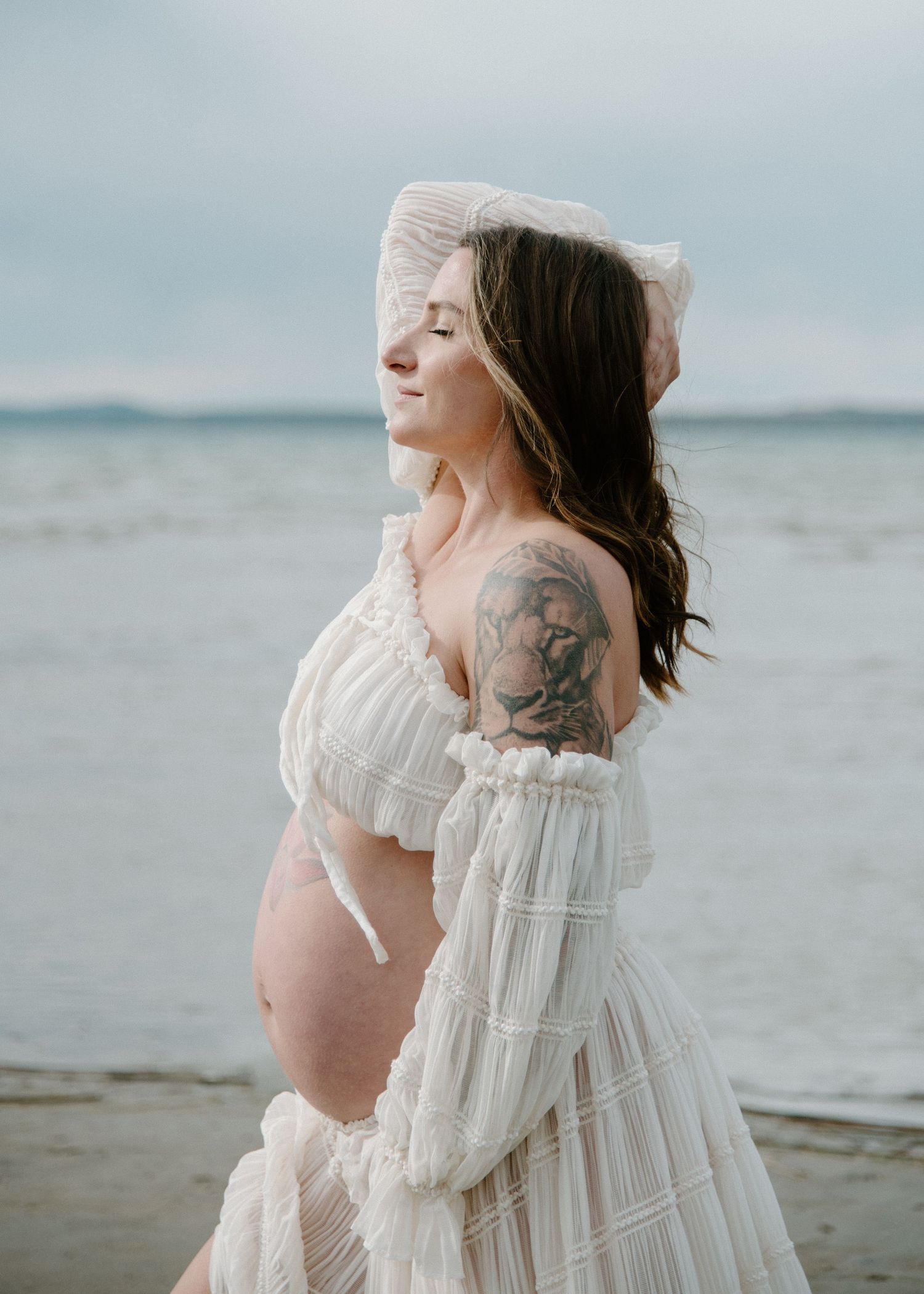 Side profile of expectant mother in white dress against serene beach backdrop. Calgary Alberta Maternity Photographer