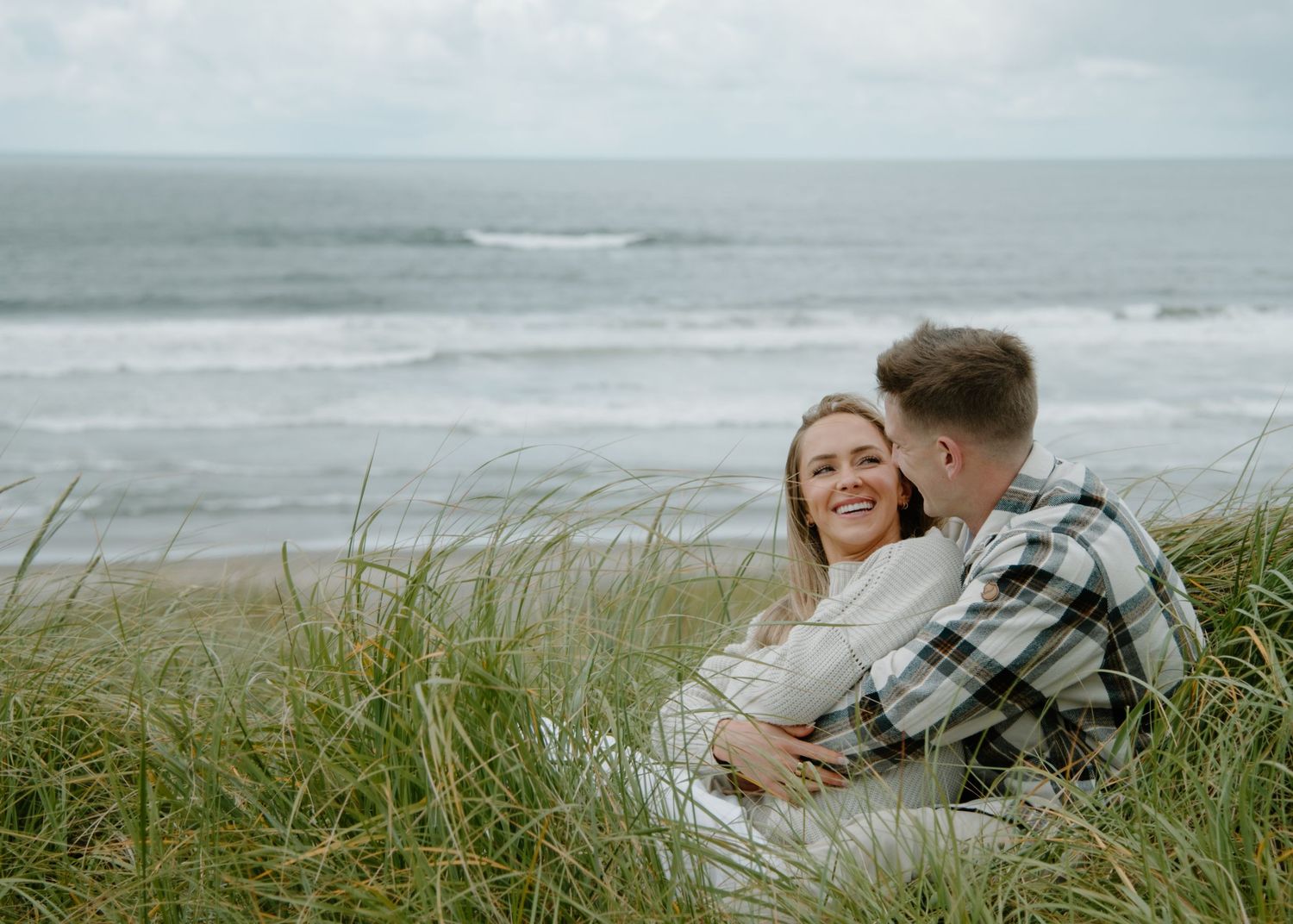 Couple sharing an intimate moment on a coastal hillside with ocean waves in background on Oregon Coast. Calgary Lifestyle and Engagement.