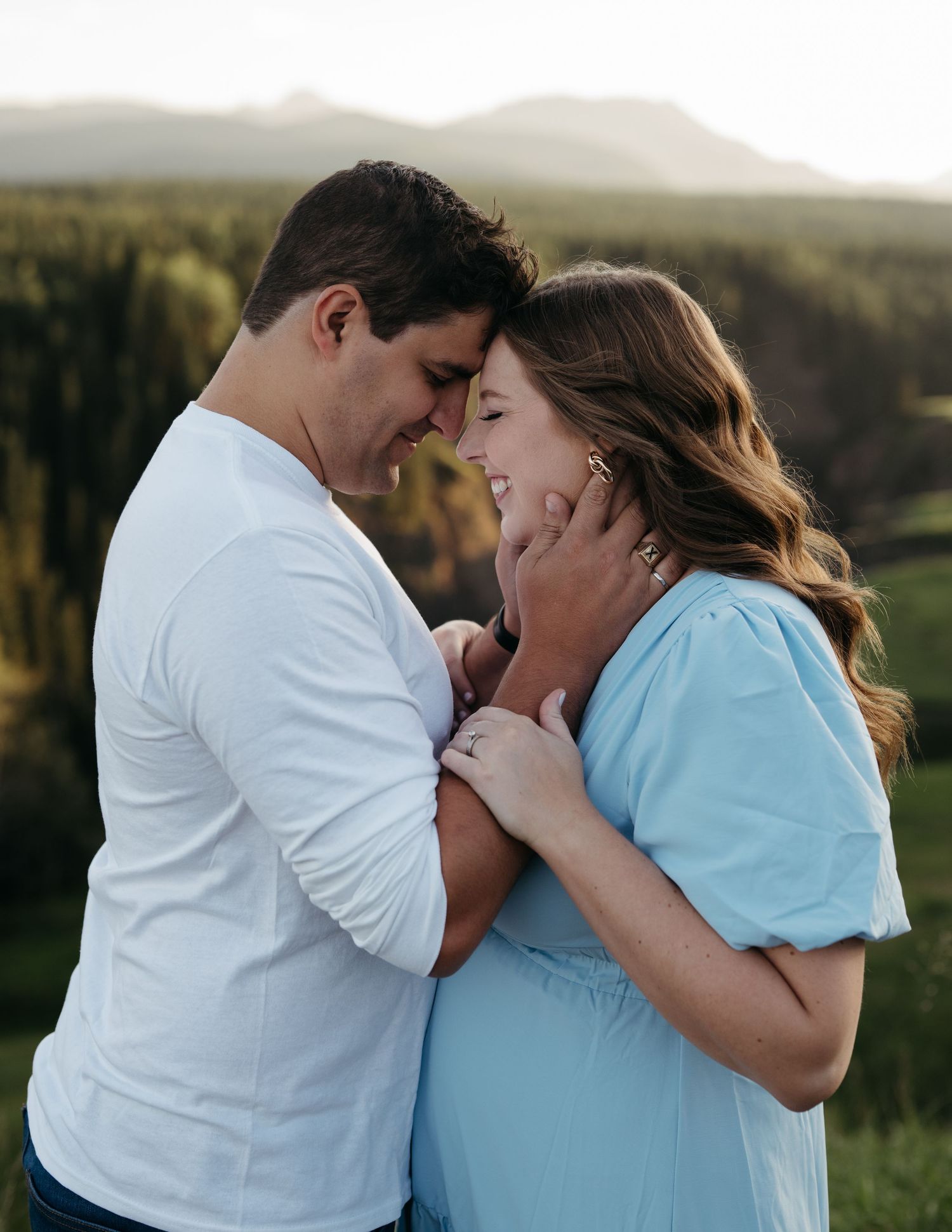 Romantic couple in white and blue outfits share an intimate moment against a scenic mountain backdrop at sunset.