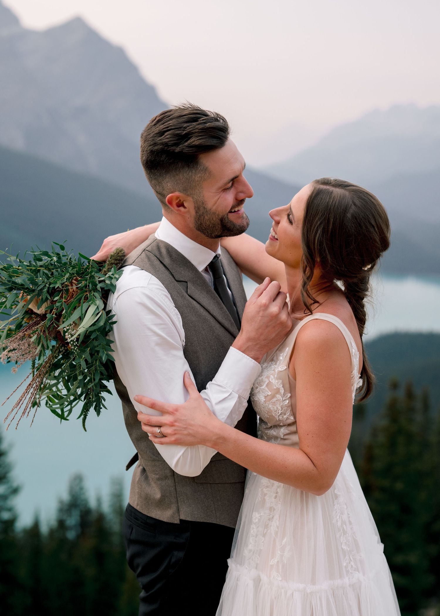 A romantic couple shares joyful moments against a misty mountain backdrop near a lake.