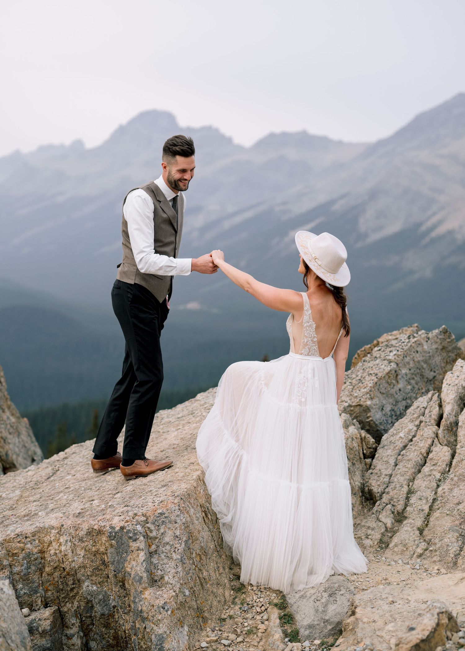 A couple shares a romantic moment atop a mountain peak with dramatic mountain ranges in the background.
