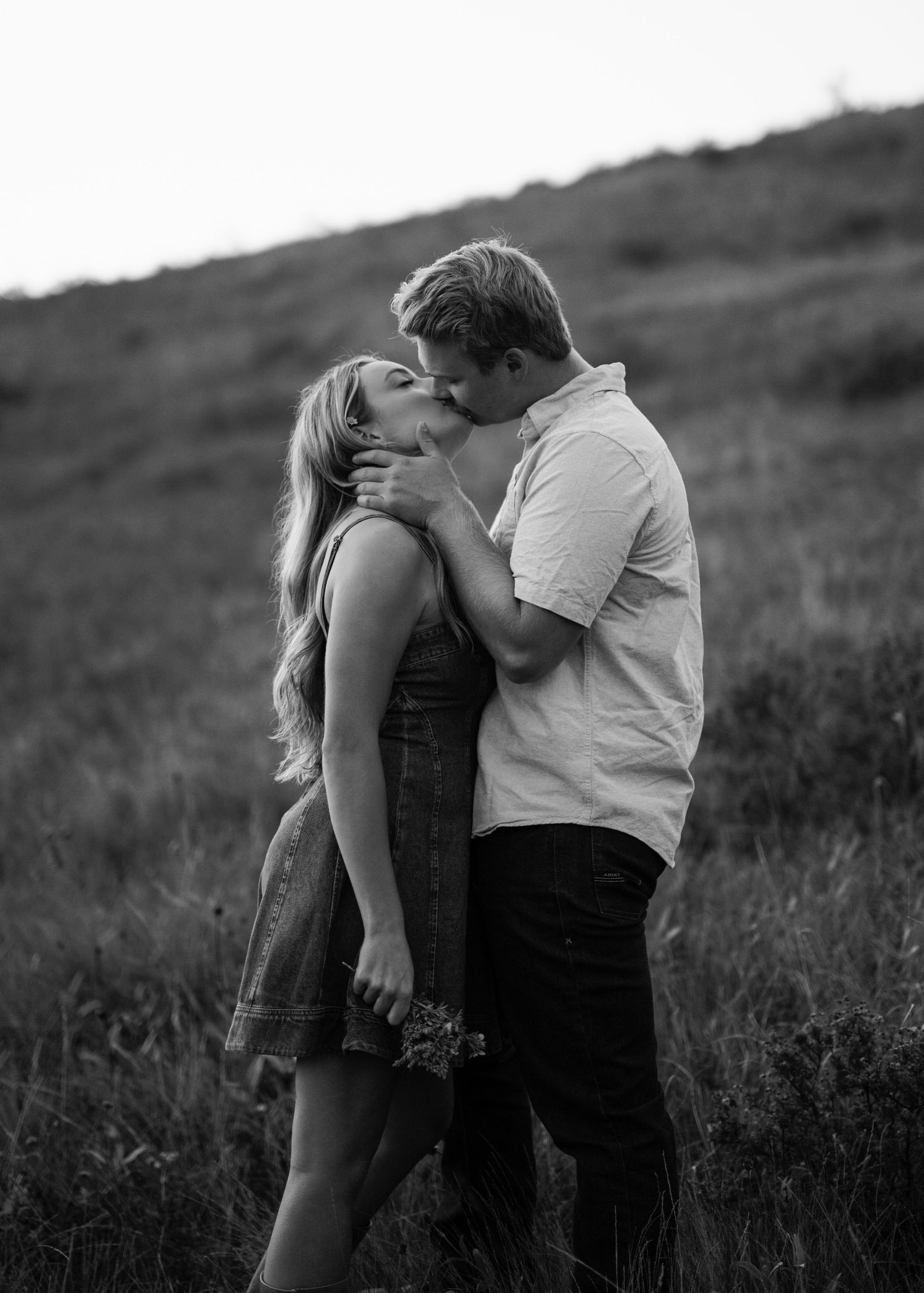 A couple shares a romantic moment in a black and white photograph taken in a grassy field.