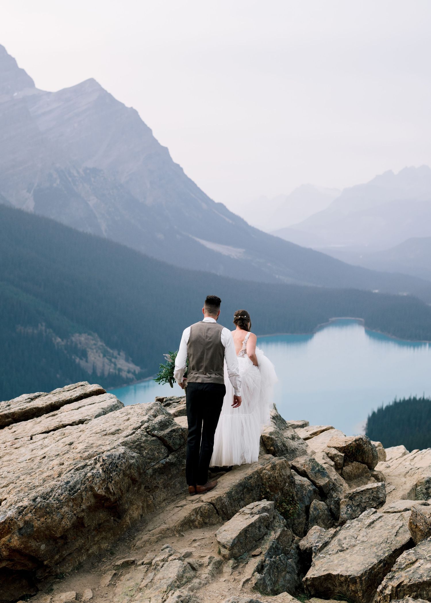 A couple stands on a rocky cliff overlooking Peyto Lake in Banff National Park with mountains in the distance.