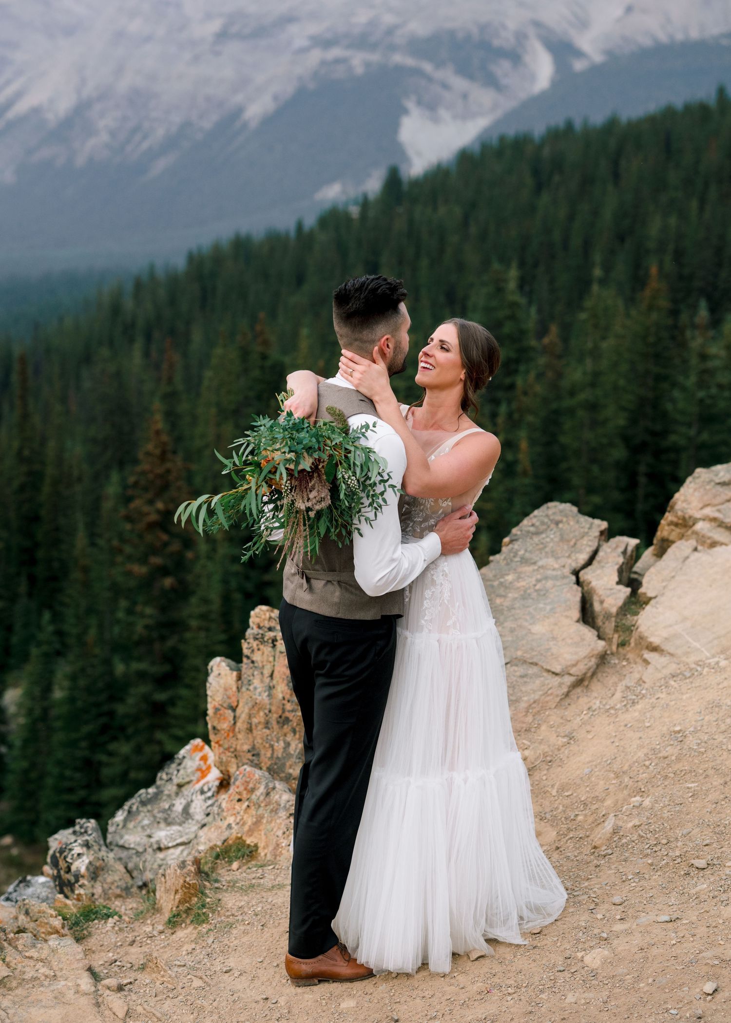 A couple in wedding attire embrace on a scenic mountain cliff overlook surrounded by evergreen forest and snow-capped peaks.