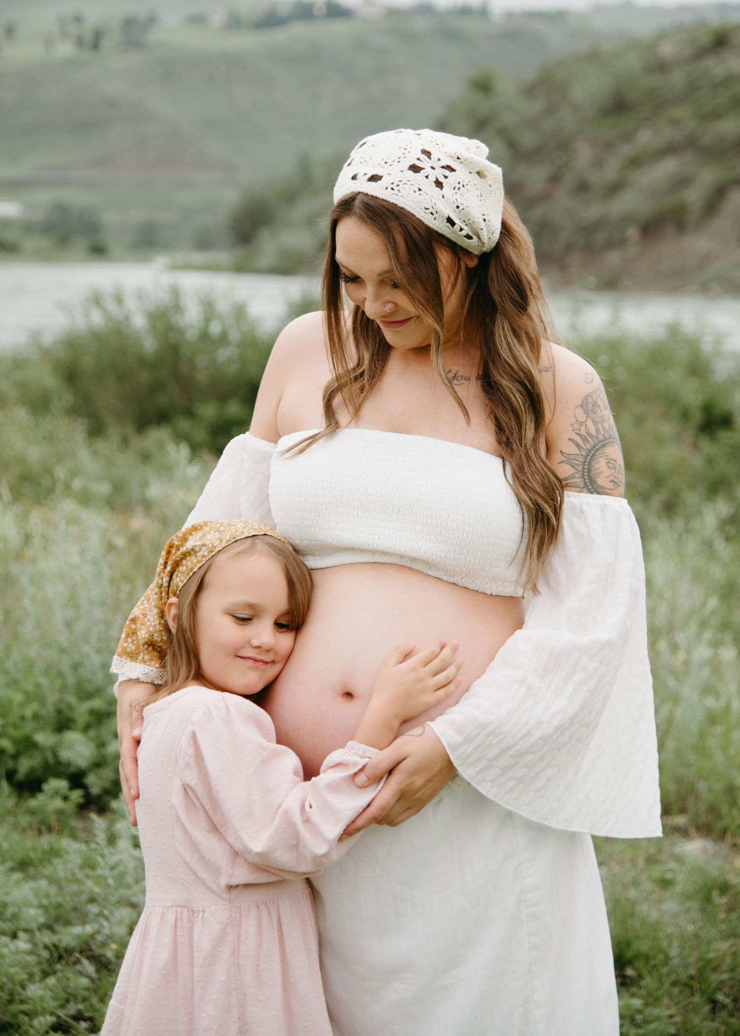 A tender maternity moment captured outdoors with white flowing dress and pink details in a natural setting.