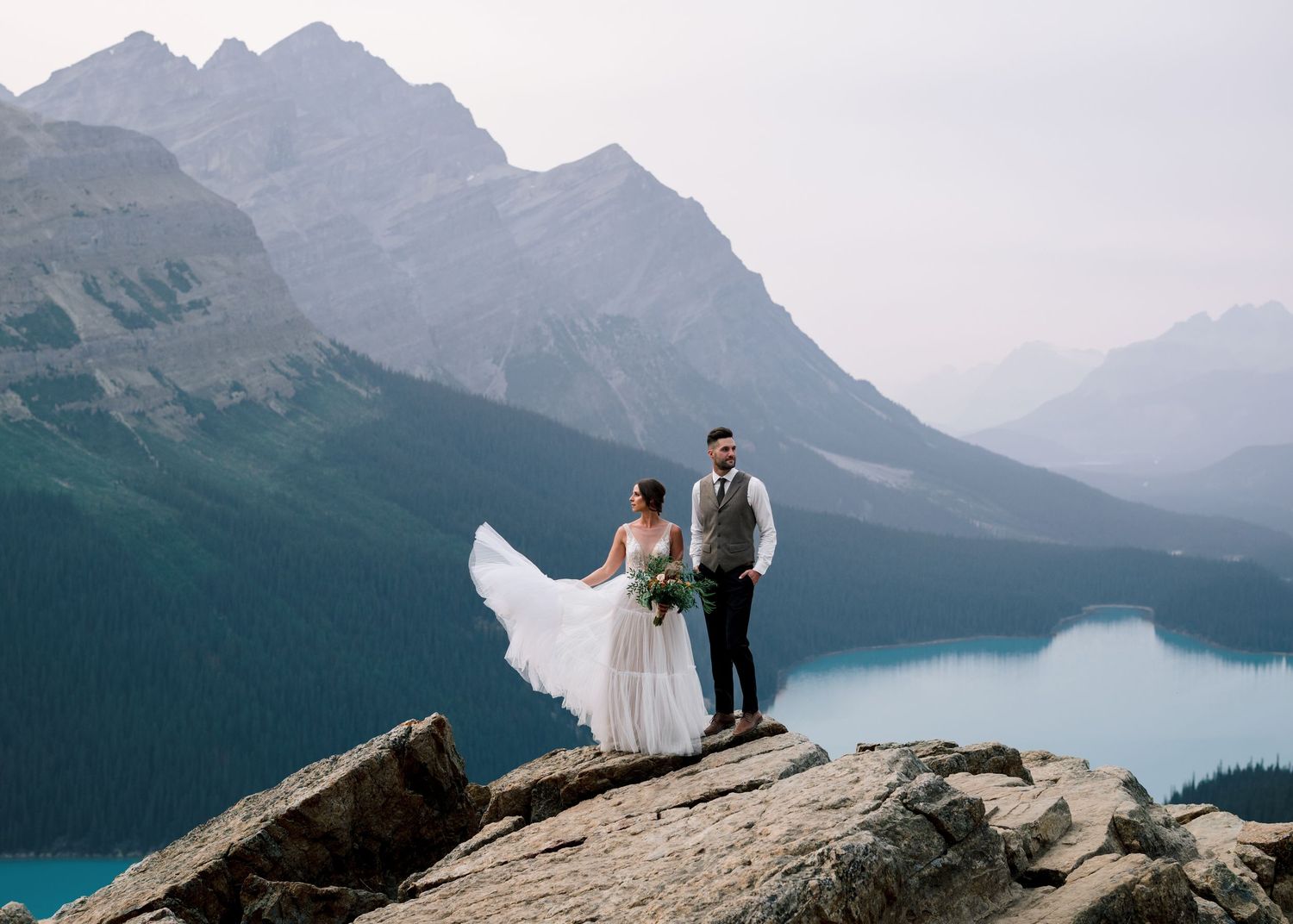 A couple stands on a rocky overlook with a stunning view of turquoise Peyto Lake and Canadian Rockies mountains.