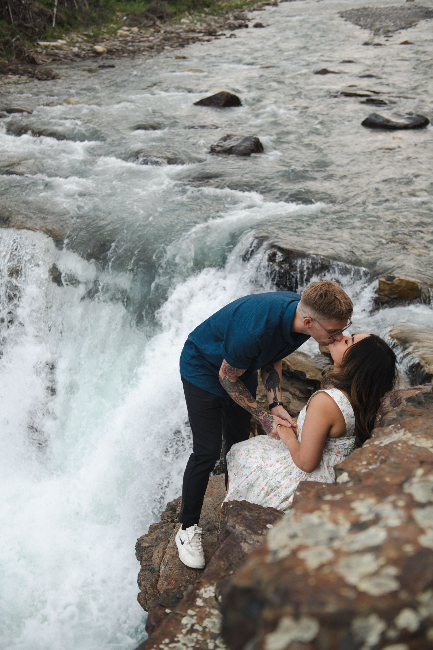 Couple sharing tender moments on cliff edge overlooking cascading waterfall in Calgary. Calgary Lifestyle and Engagement Photographer.