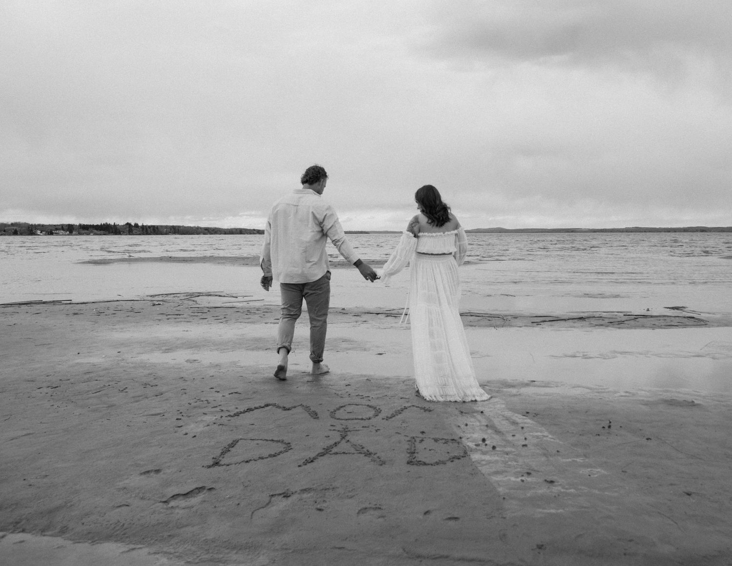 A couple walking hand in hand on beach with message written in sand. Calgary Alberta Maternity Photographer.