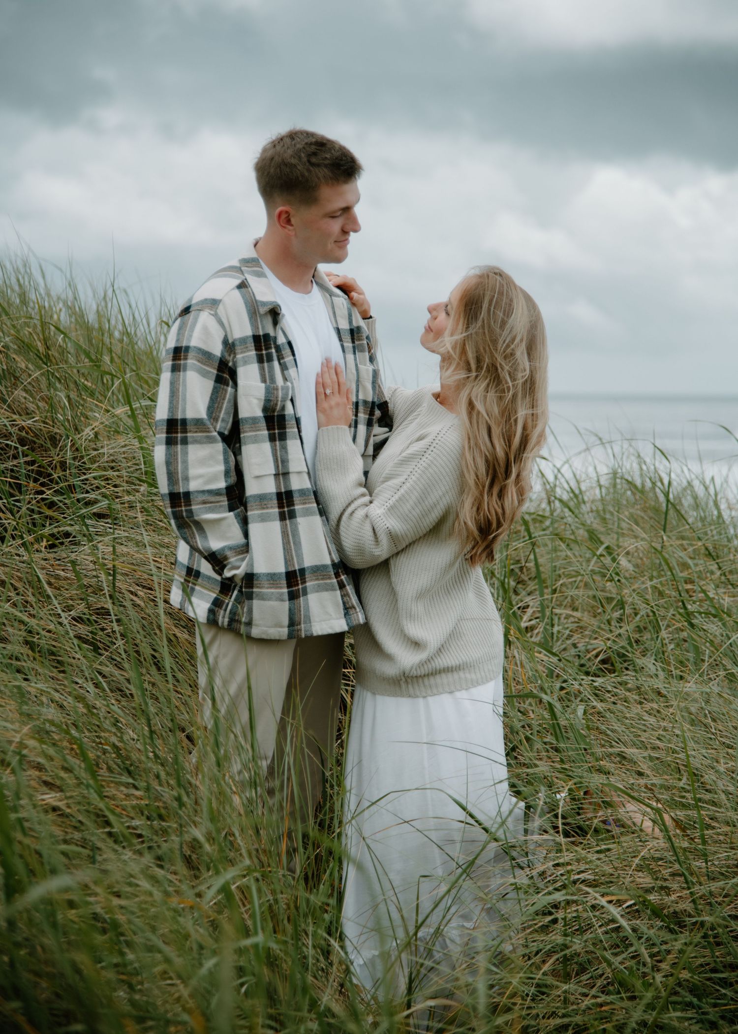 Couple in coordinating outfits share tender moment among windswept coastal grasses on Oregon Coast. Calgary Lifestyle and Engagement.