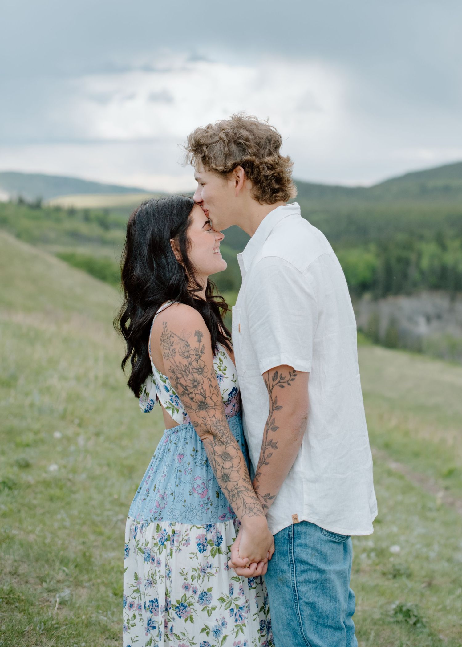 A romantic couple standing on a grassy hillside with mountains in the background wearing white and floral print. Calgary Alberta Engagement