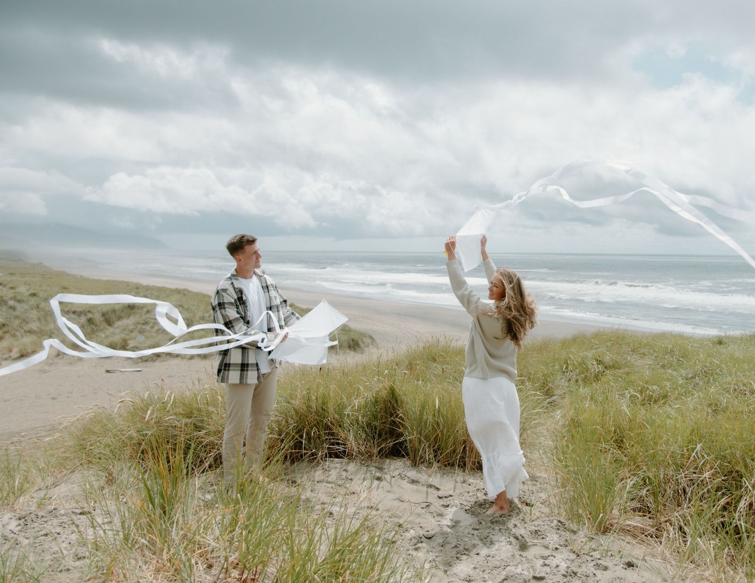 Two people playfully toss white ribbons in the air on a scenic beach hill. Calgary Alberta Engagement Photographer.