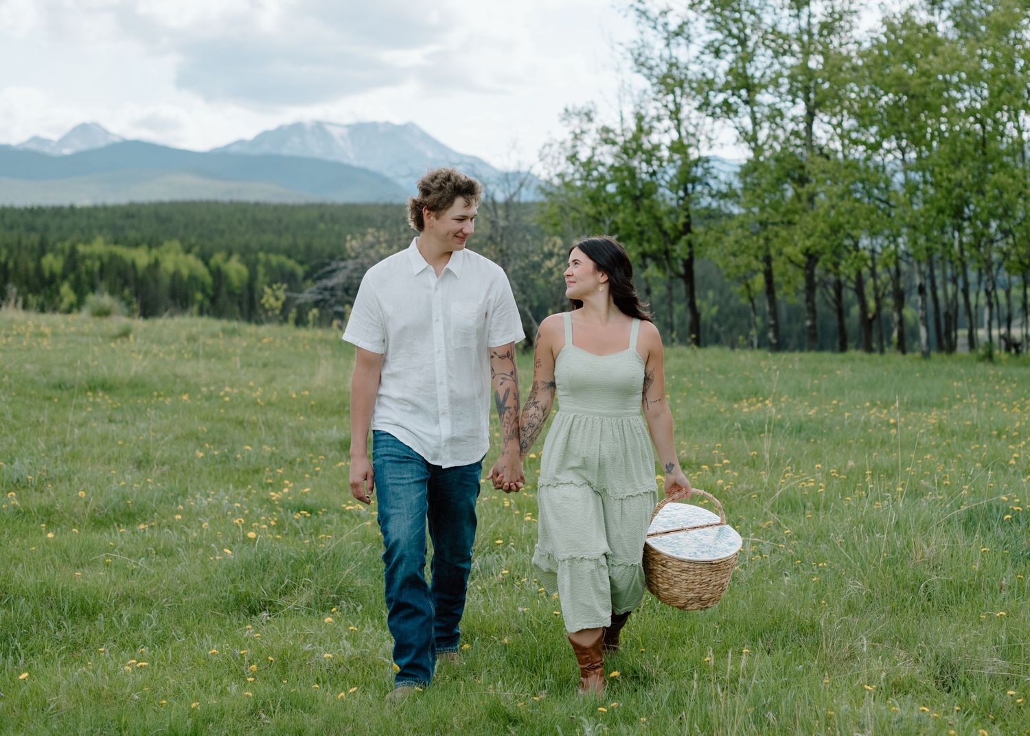 A couple strolls through a grassy meadow carrying a picnic basket with mountains in the background. Calgary Alberta Engagement Photographer.