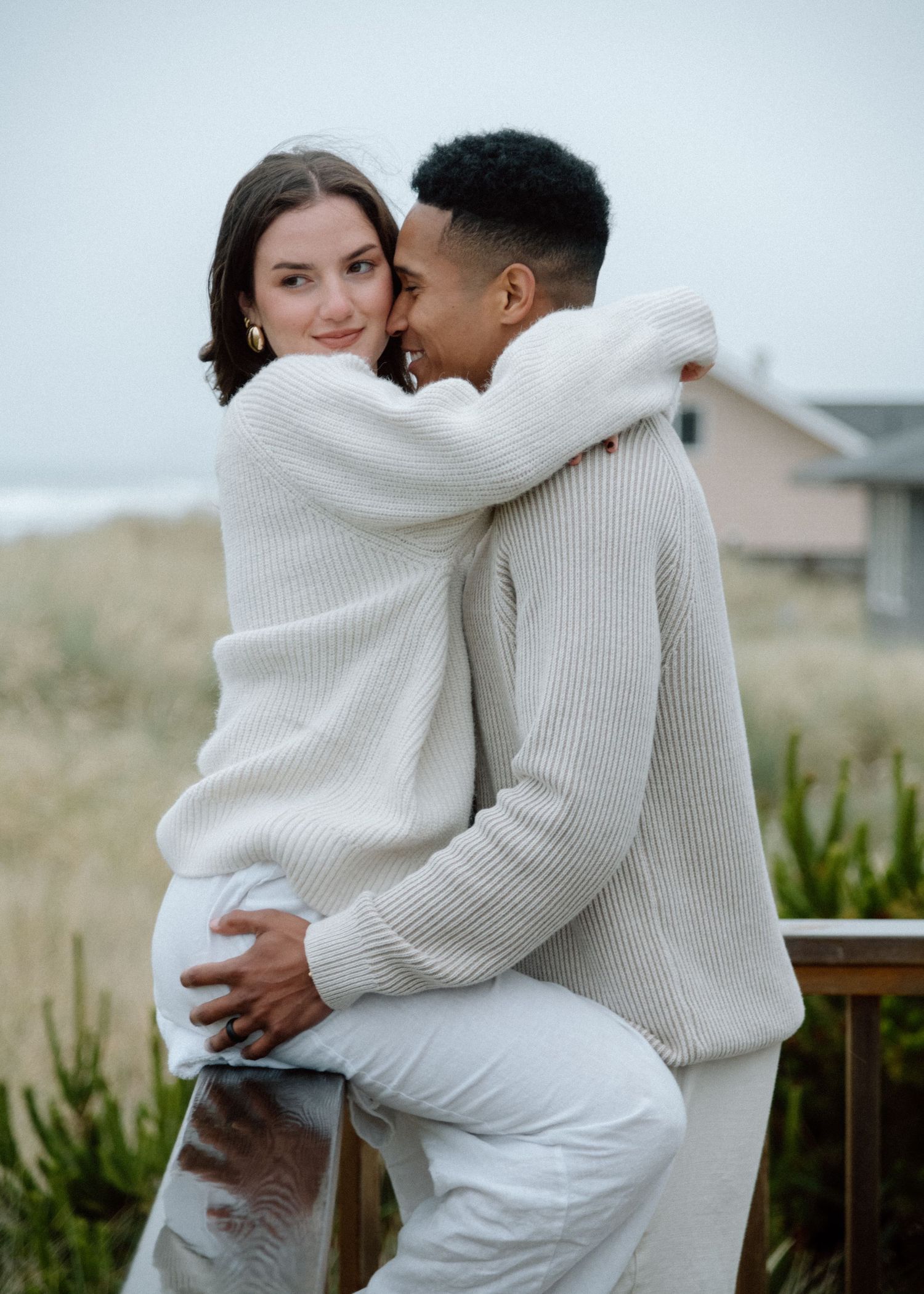 Romantic couple in matching cream sweaters embrace each other on a cloudy coastal day.