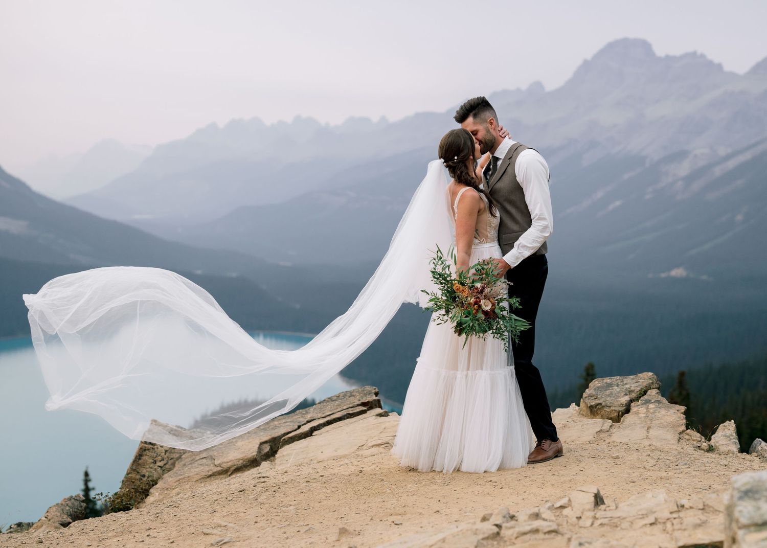 A wedding couple shares an intimate moment against a stunning mountain backdrop with a flowing white veil in the wind.