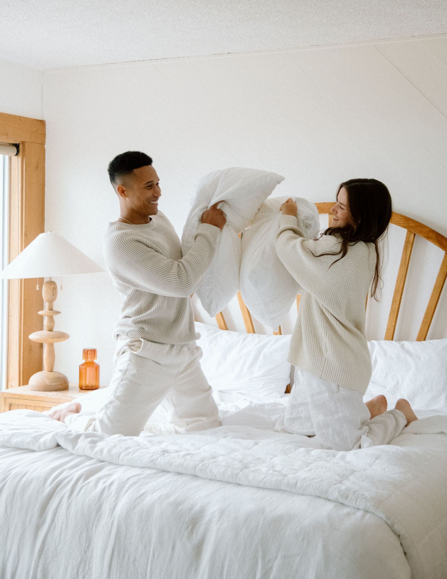 Two people in white outfits having a playful pillow fight on a cozy bed with white linens and wood furniture.
