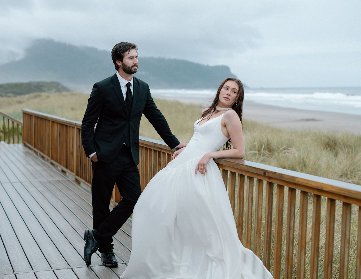 A couple in wedding attire shares a joyful moment on a wooden boardwalk overlooking a misty beach.