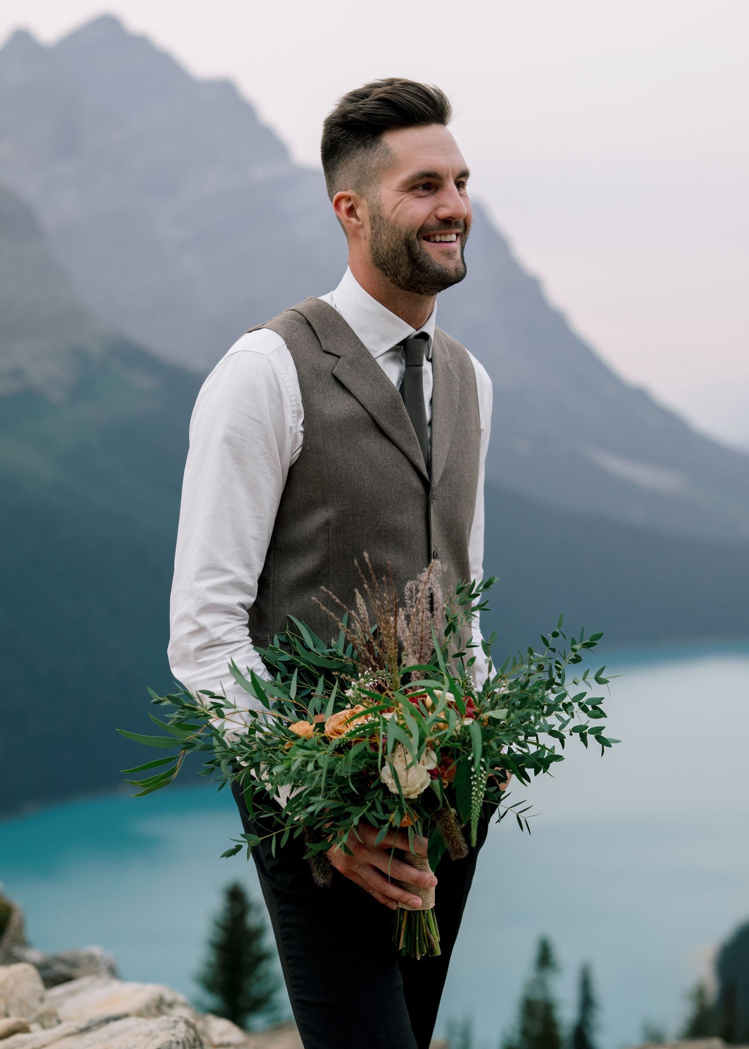 Man in formal vest and tie smiles outdoors with mountain lake and evergreen trees in background holding floral greenery.