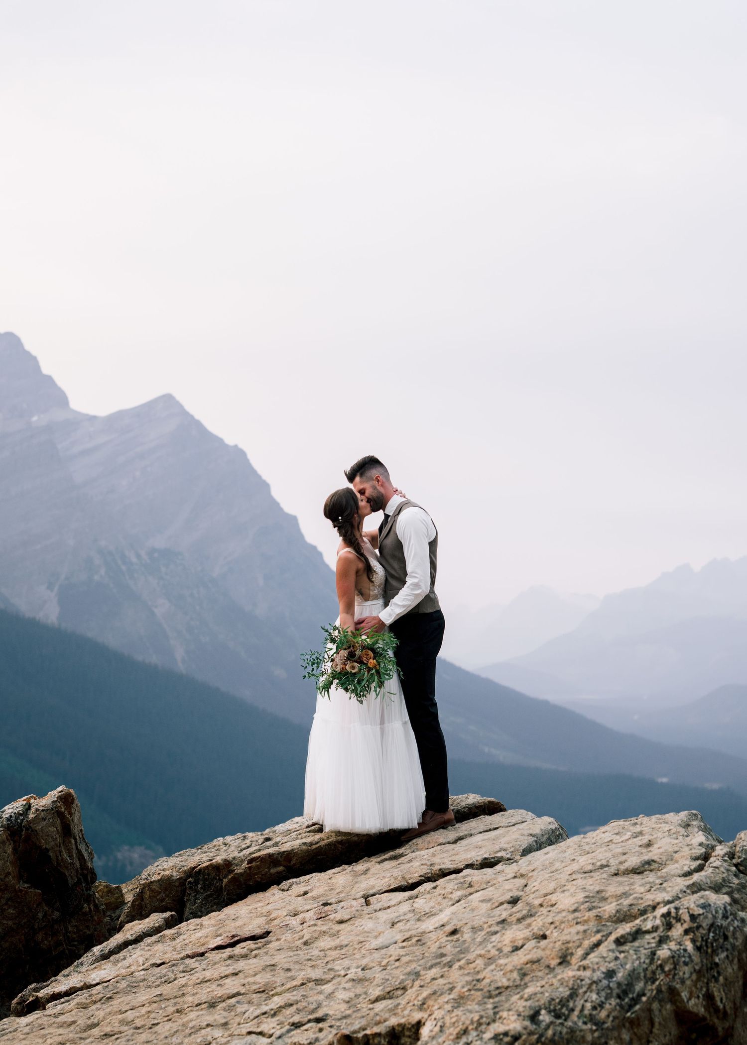 Wedding couple embraces on a rocky mountain outcrop with dramatic mountain peaks and valleys in the background.