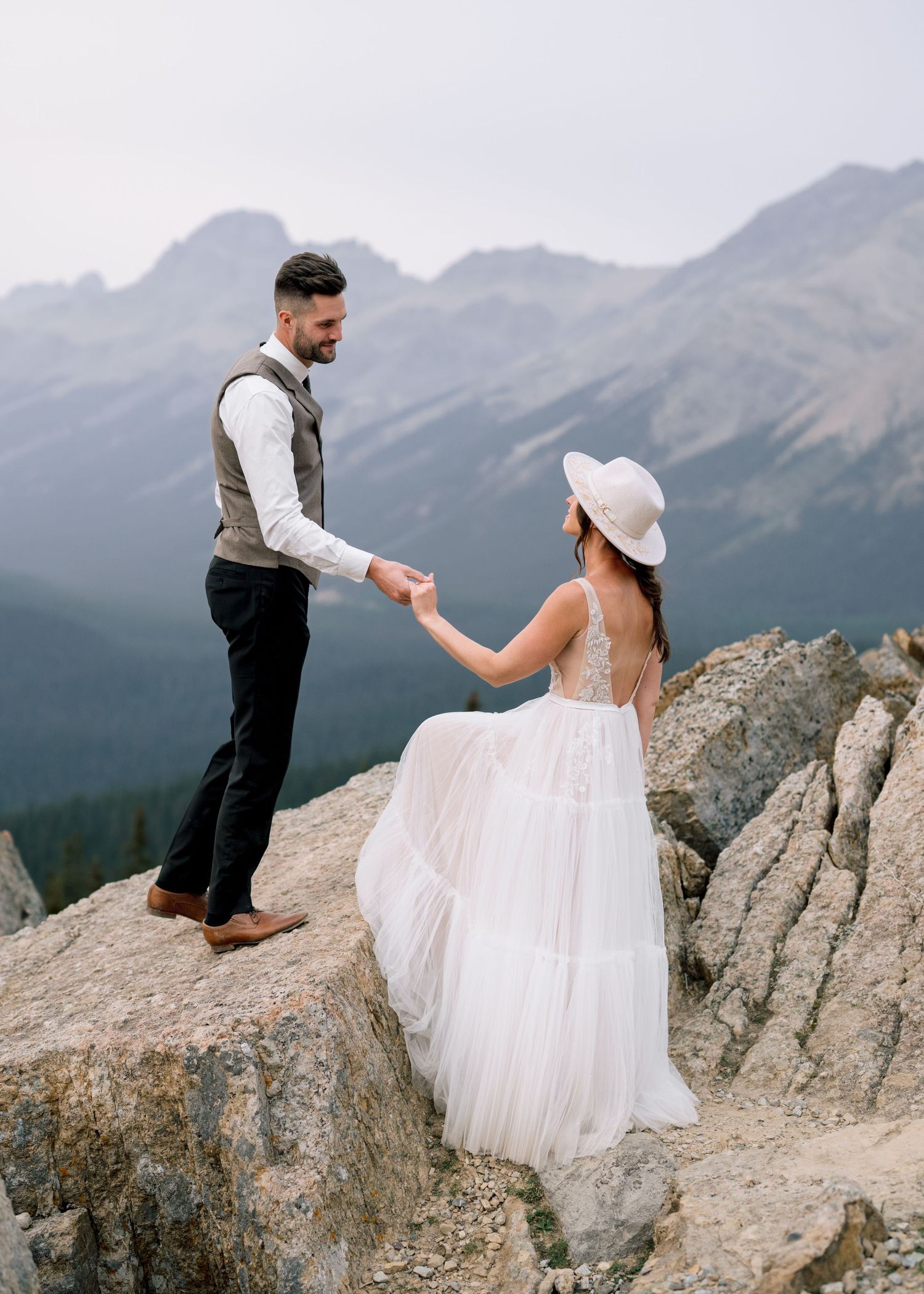 A couple shares a romantic moment on a rocky mountain overlook with dramatic mountain peaks in the background.
