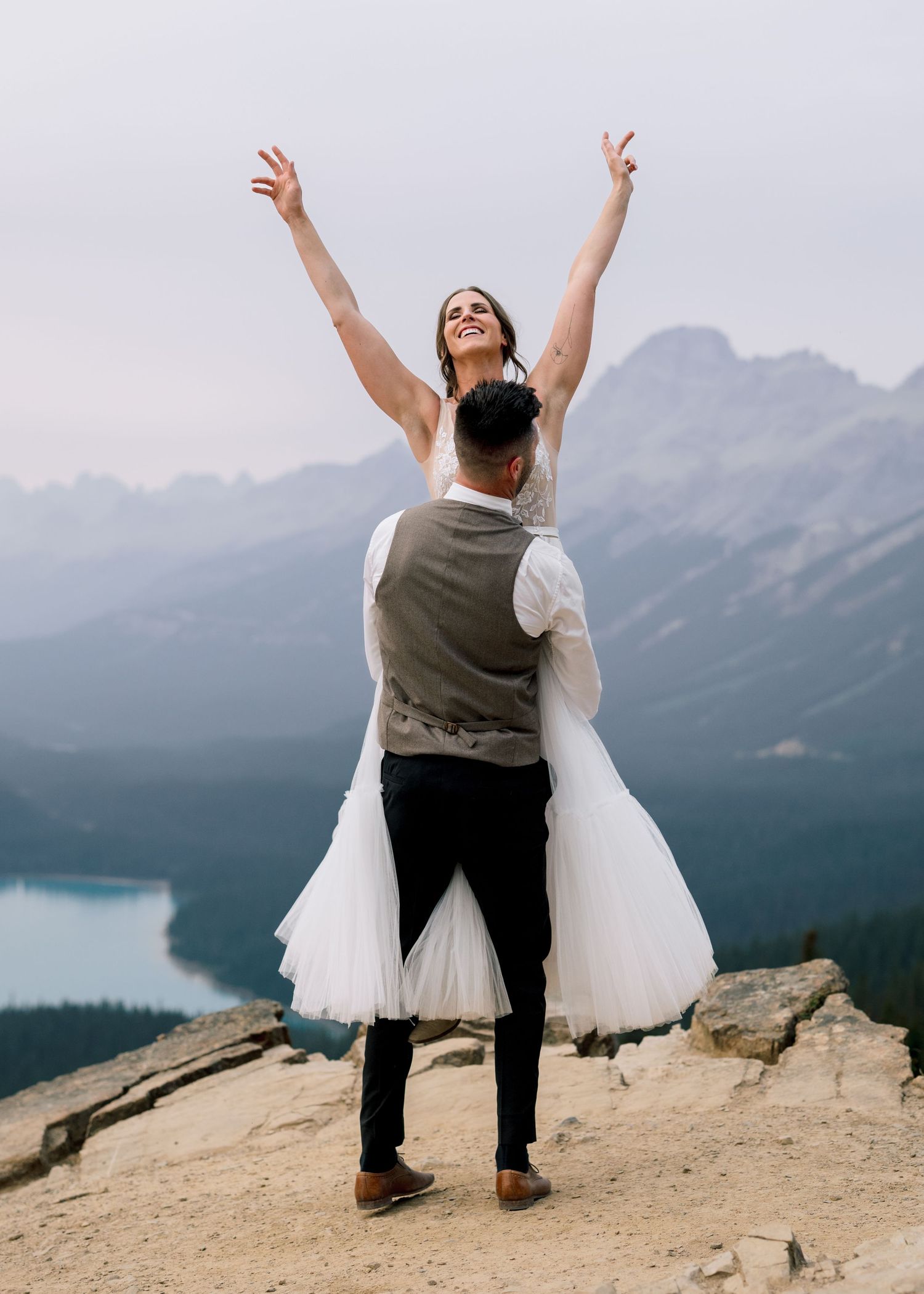 Two people in wedding attire celebrate on a mountaintop overlooking a scenic mountain lake in Banff National Park.