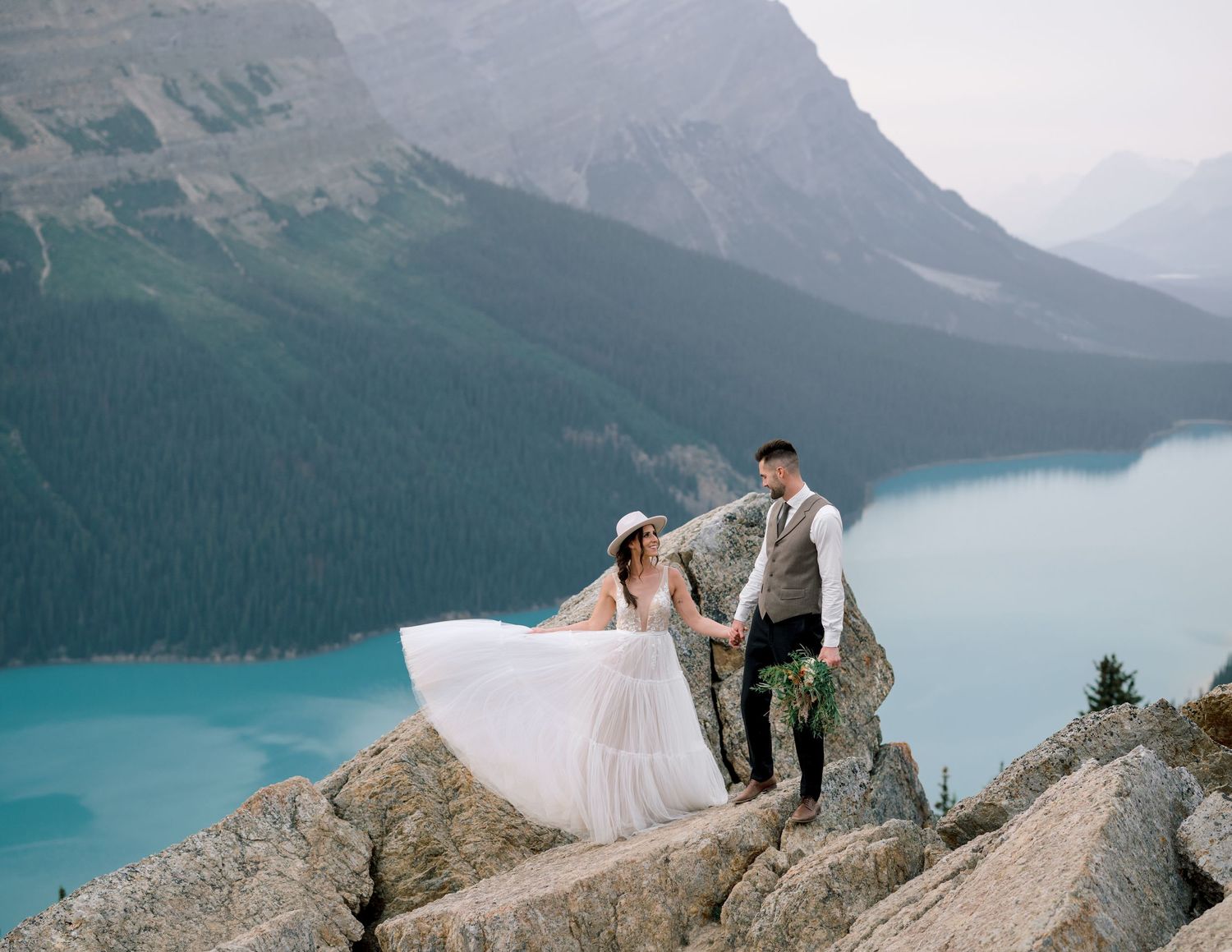 A couple in wedding attire stands together on a rocky cliff overlooking the turquoise waters of Peyto Lake in Banff National Park.