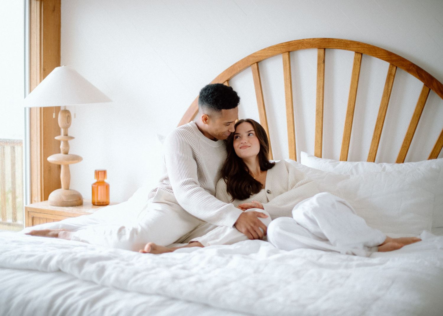 A romantic series of lifestyle photos showing a couple lounging together in a bright white bed near windows.